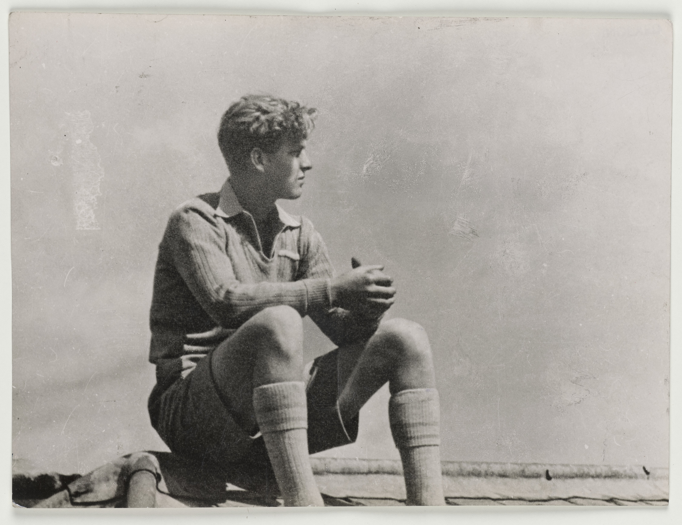 Full length, informal portrait of Prince Philip sitting on the roof of Gordonstoun School, facing right side profile. He wears school uniform. This image reproduced in Five Gold Rings, published in 2007.