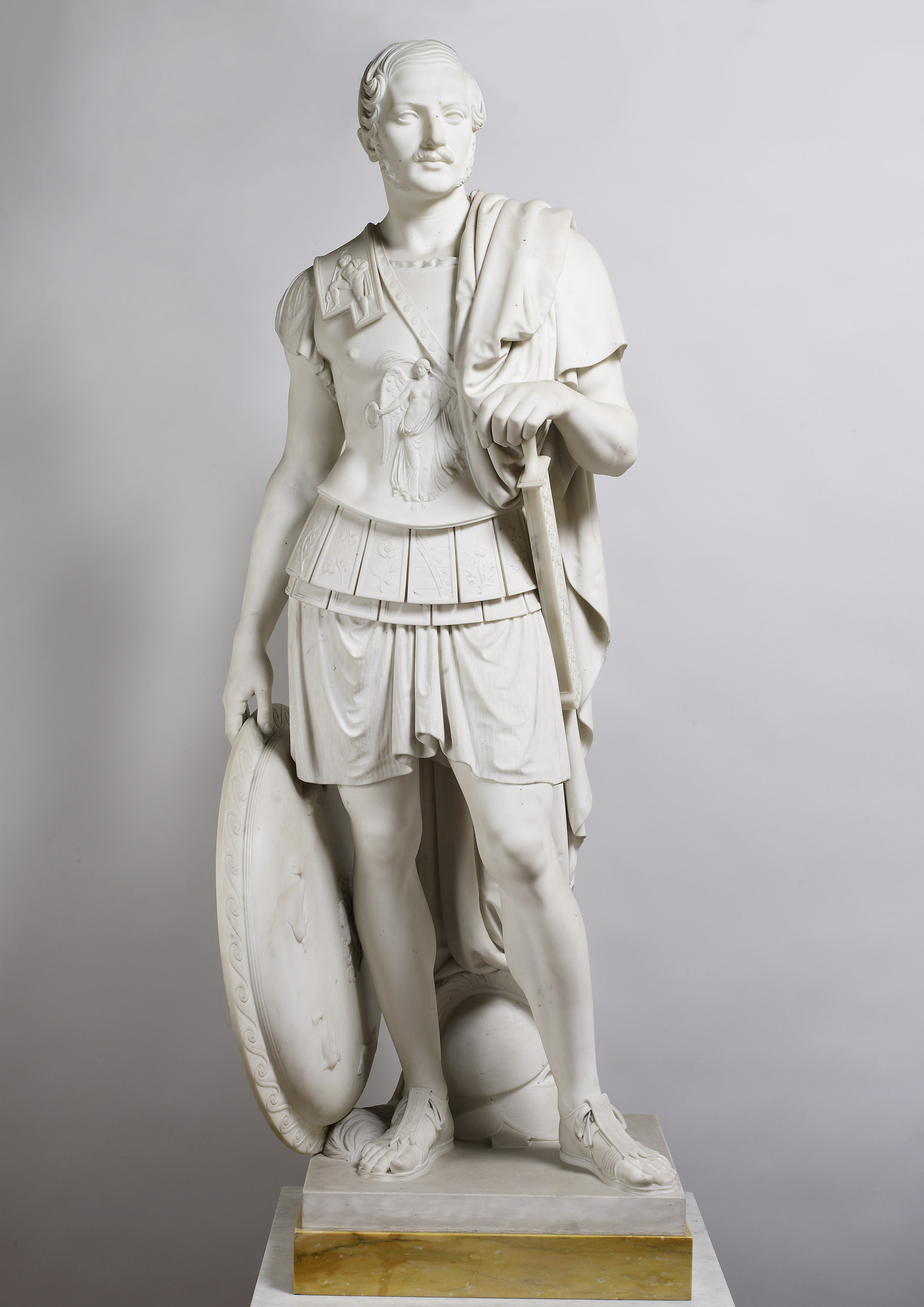 Prince Albert (1819-1861) Full length marble statue of Prince Albert wearing a classical suit of armour chased with emblems of the British Isles and Coburg, with a cloak over his left shoulder. His left hand rests on the hilt of his sheathed sword; his right holds a circular shiel