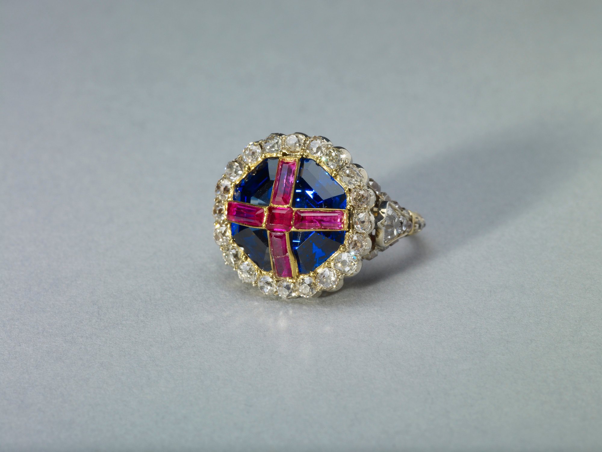 The ring comprises an octagonal step-cut sapphire, open-set in gold, overlaid with four oblong and one square rubies in gold strips forming a cross, within a border of twenty cushion-shaped brilliants in transparent silver collets. Brilliants decorate the