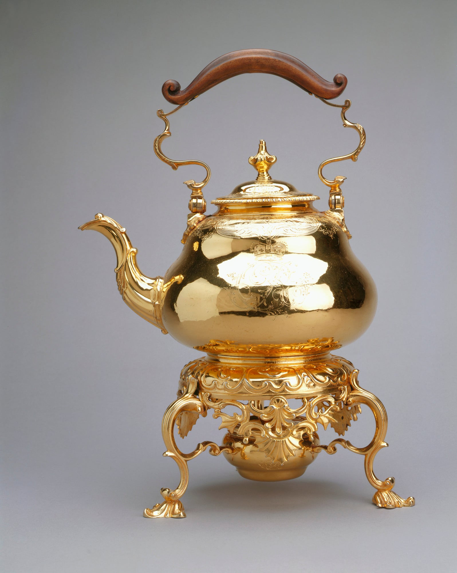 Silver gilt tea kettle. Hinged bail handle with ebonised wooden grip. Domed detachable cover with baluster finial. Pear shaped kettle with curved spout; engraved with foliate scrolls and crowned CR monogram. Circular stand with pierced apron; on 3 scroll 