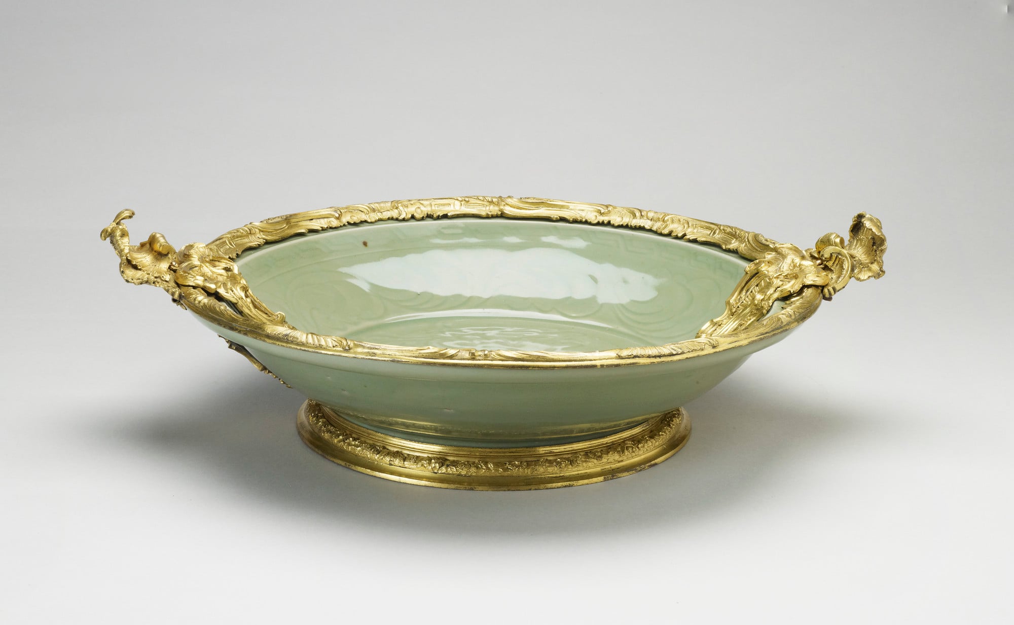 Dish Chinese porcellanous stoneware dish, with carved decoration under a thick grey-green celadon glaze and French gilt-bronze mounts
Thickly potted, with narrow, spreading rim. Carved in the centre is a flower spray and round the sides a scrolling design, wi