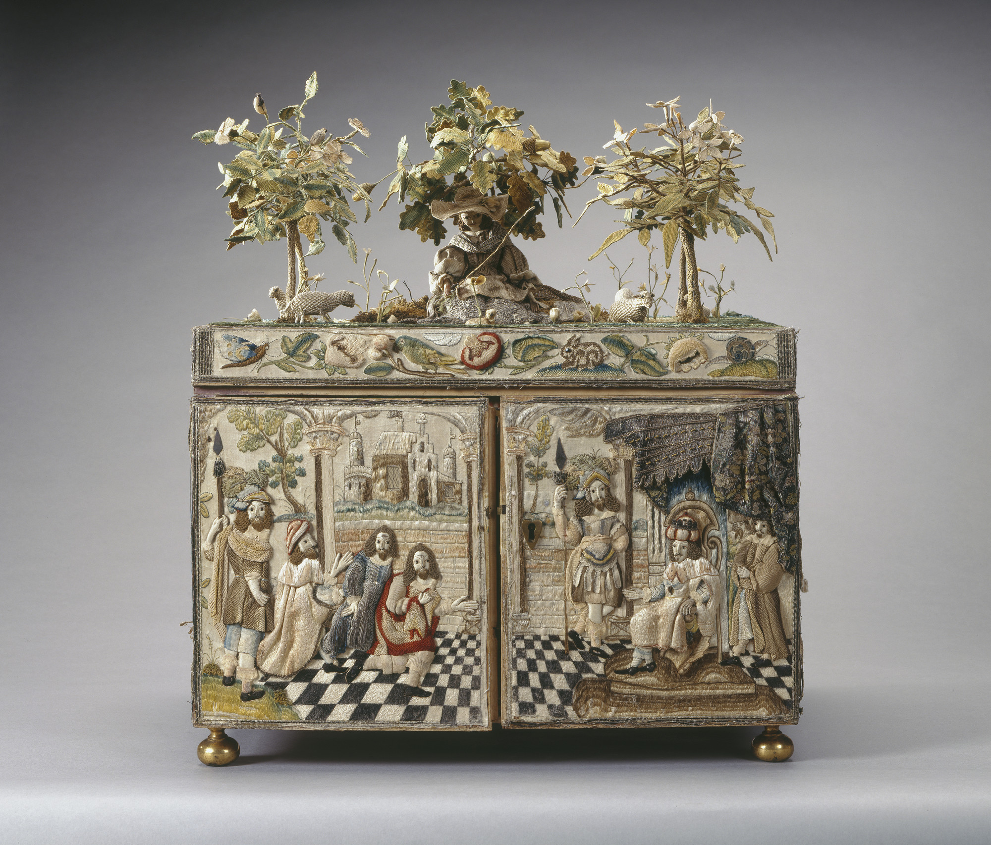 A stumpwork casket with two doors and brass drop handles and interior fitted with mirror and small drawers.Top decorated with sheperdess wearing a hat with four sheep, a lamb and a dog, sitting under an oak tree.
Embroidered caskets decorated with raised 