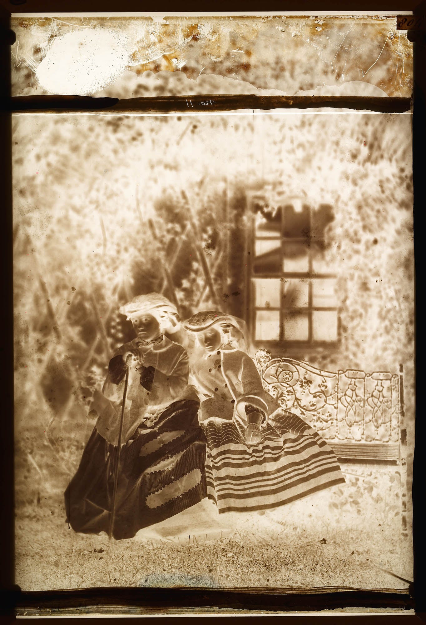 Glass plate negative of a full length portrait of Victoria, Princess&nbsp;Royal&nbsp;and Princess Alice. The sisters&nbsp;are seated together on a decorative ironwork garden seat. The Princess Royal holds a walking stick with Princess Alice leaning on her