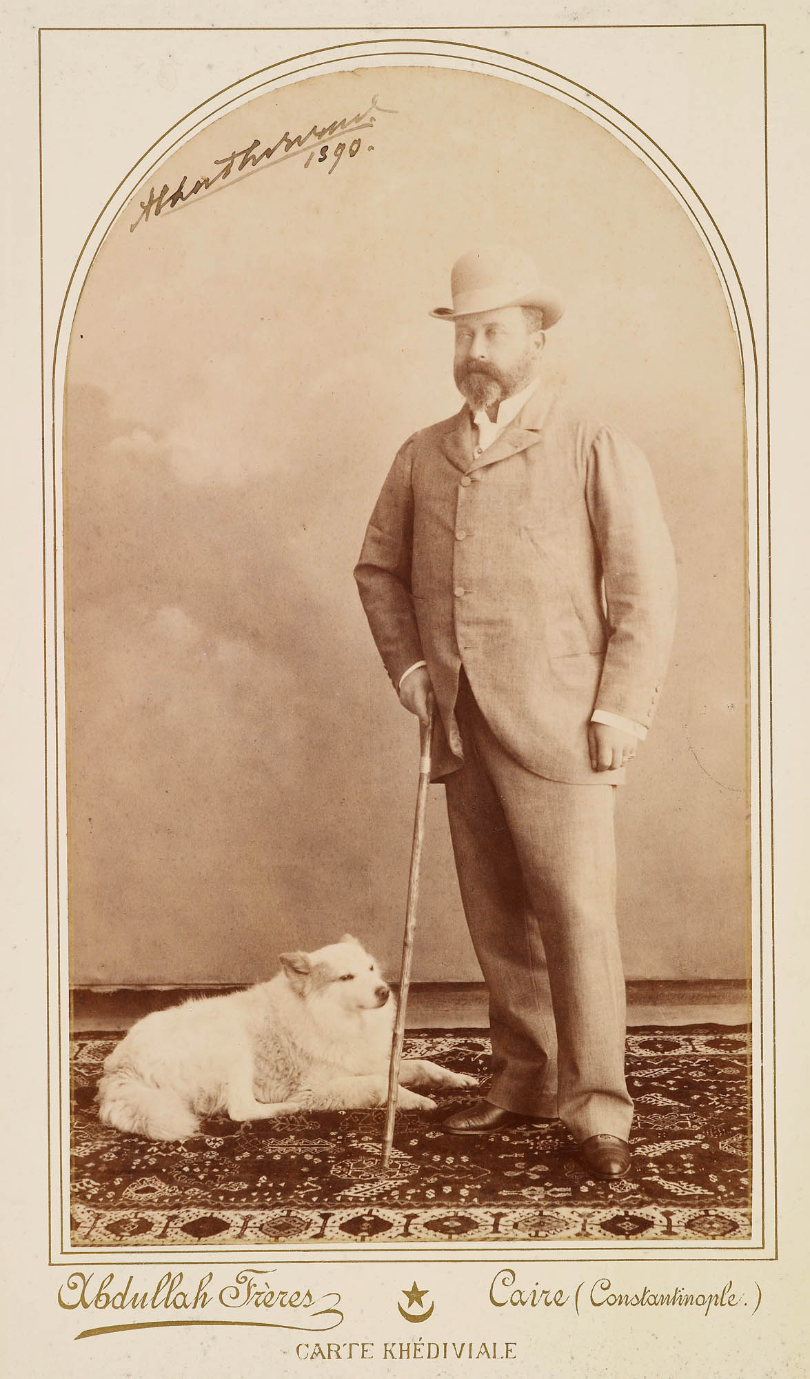 Full-length portrait photograph of Albert Edward, Prince of Wales, later King Edward VII wearing a suit and bowler hat, holding a walking stick in his right hand, standing on a rug with a white dog, probably a Samoyed, laying on the rug to the Prince's ri