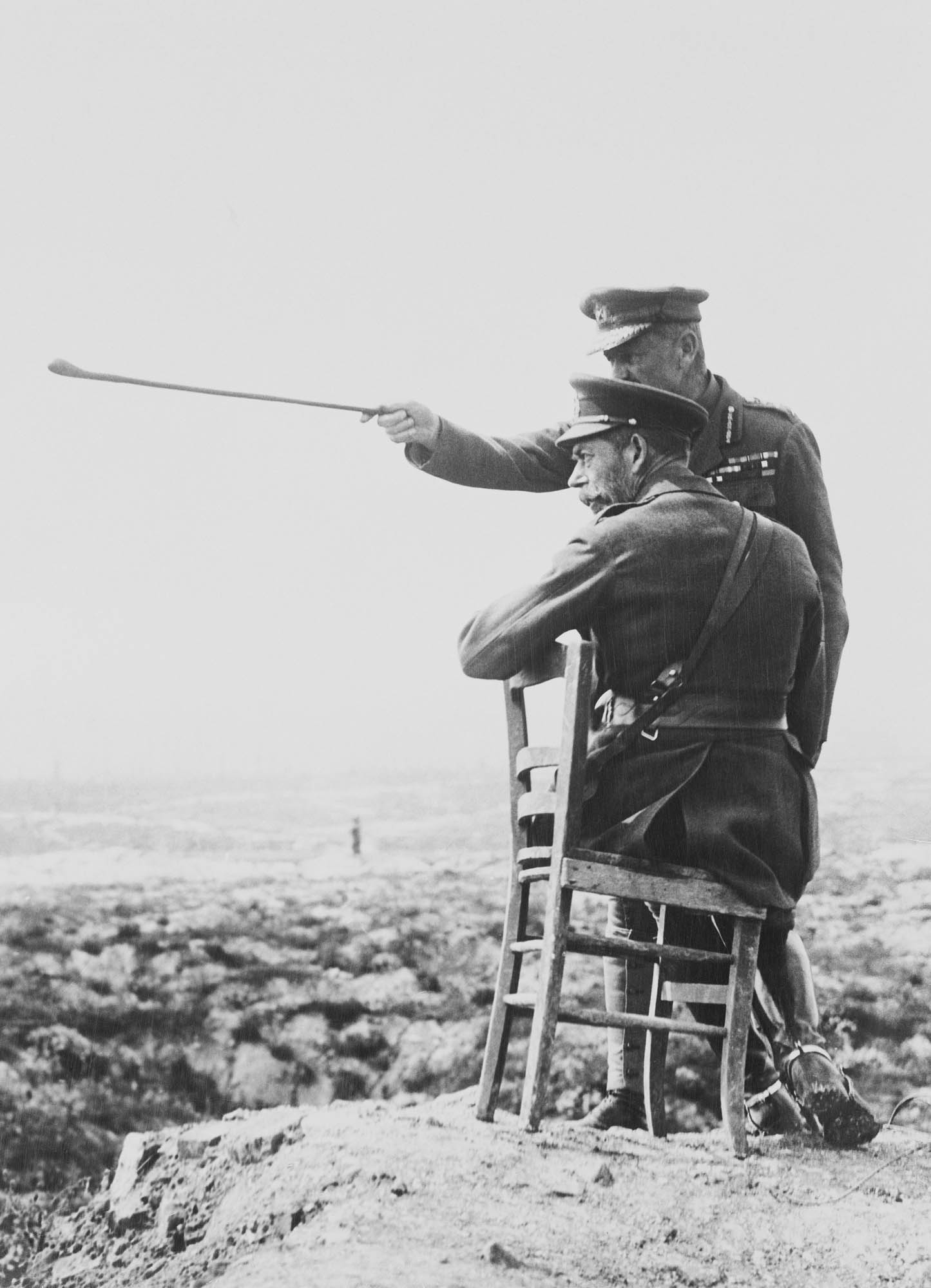 Photograph of King George V with Officers during the First World War, c. 1914-1918 Photograph showing King George V (1865-1936) sitting on a chair on top of a spoil heap, talking with an unidentified gentleman wearing military uniform who is pointing his stick out, which the King is looking towards in the distance. There is a derelict l