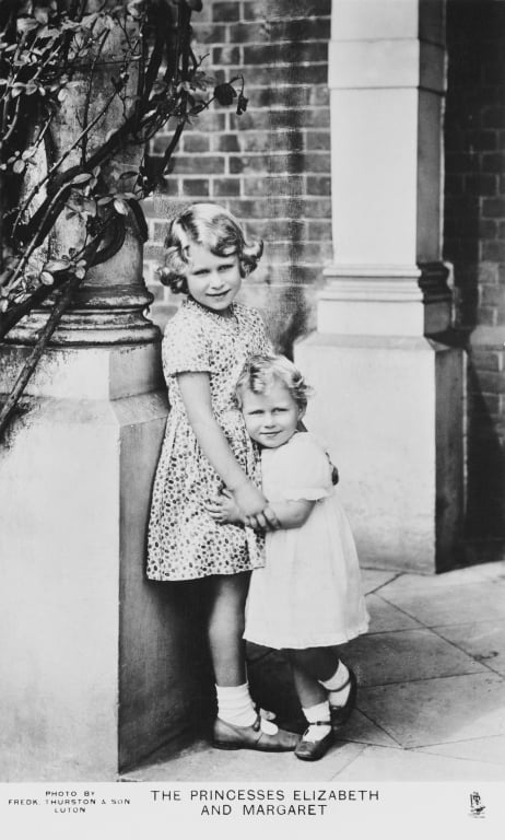 Double portrait post card photograph of HM Queen Elizabeth II (1926- ) when Princess Elizabeth of York and Princess Margaret (1930-2002) when Princess Margaret of York, standing together next to a pillar outside an unknown property