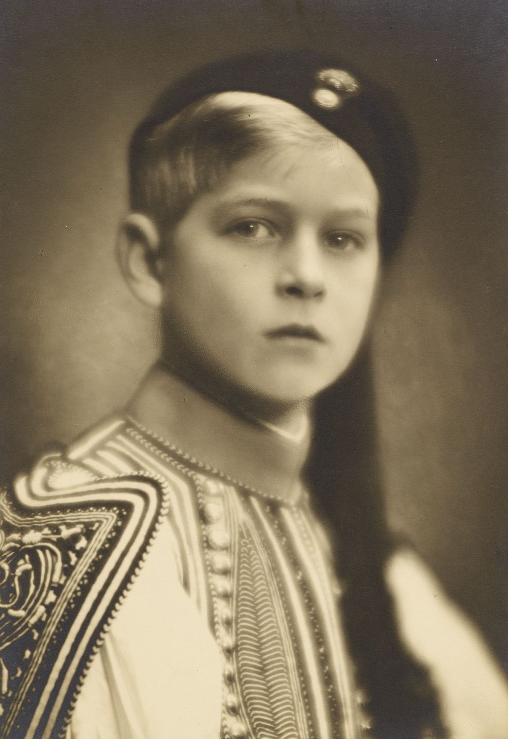Photograph of a head and shoulders length portrait of Prince Philip of Greece, later HRH The Duke of Edinburgh, facing the viewer. He wears traditional Greek costume and hat.