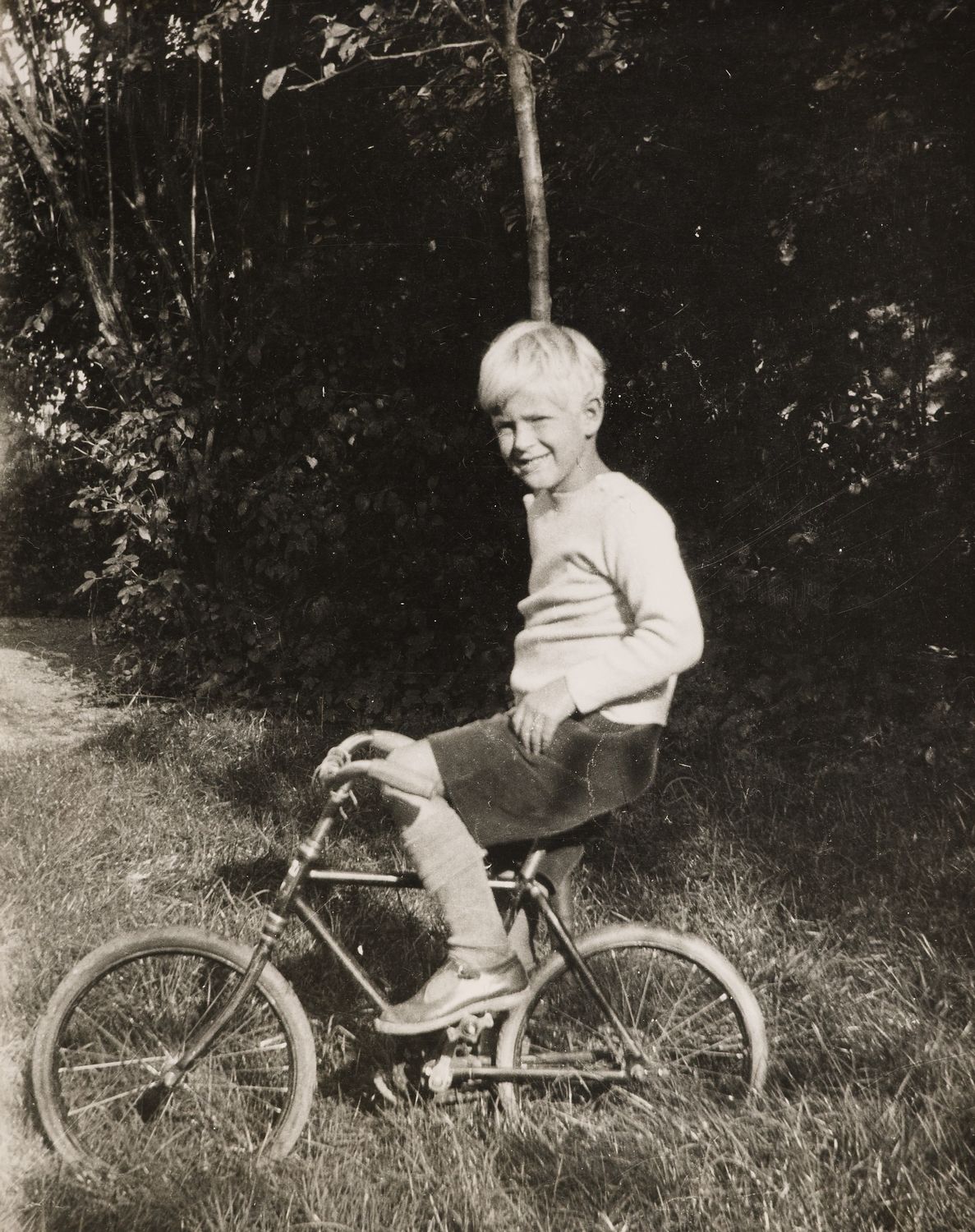 Photograph of Prince Philip of Greece, later HRH The Duke of Edinburgh, seated on a bicycle in left side profile. He faces the viewer, smiling.