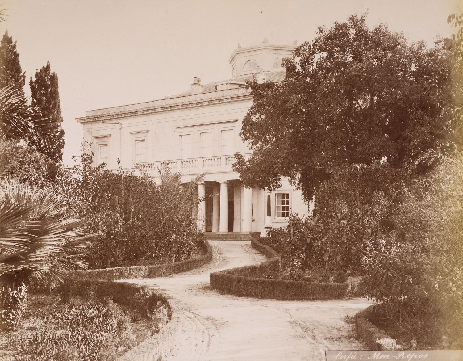 Photograph of Mon Repos villa on Corfu with a view up chicane in driveway, between low hedges separating it from beds of shrubs and trees. A neo-classical house stands at the end with a colonnaded porch.