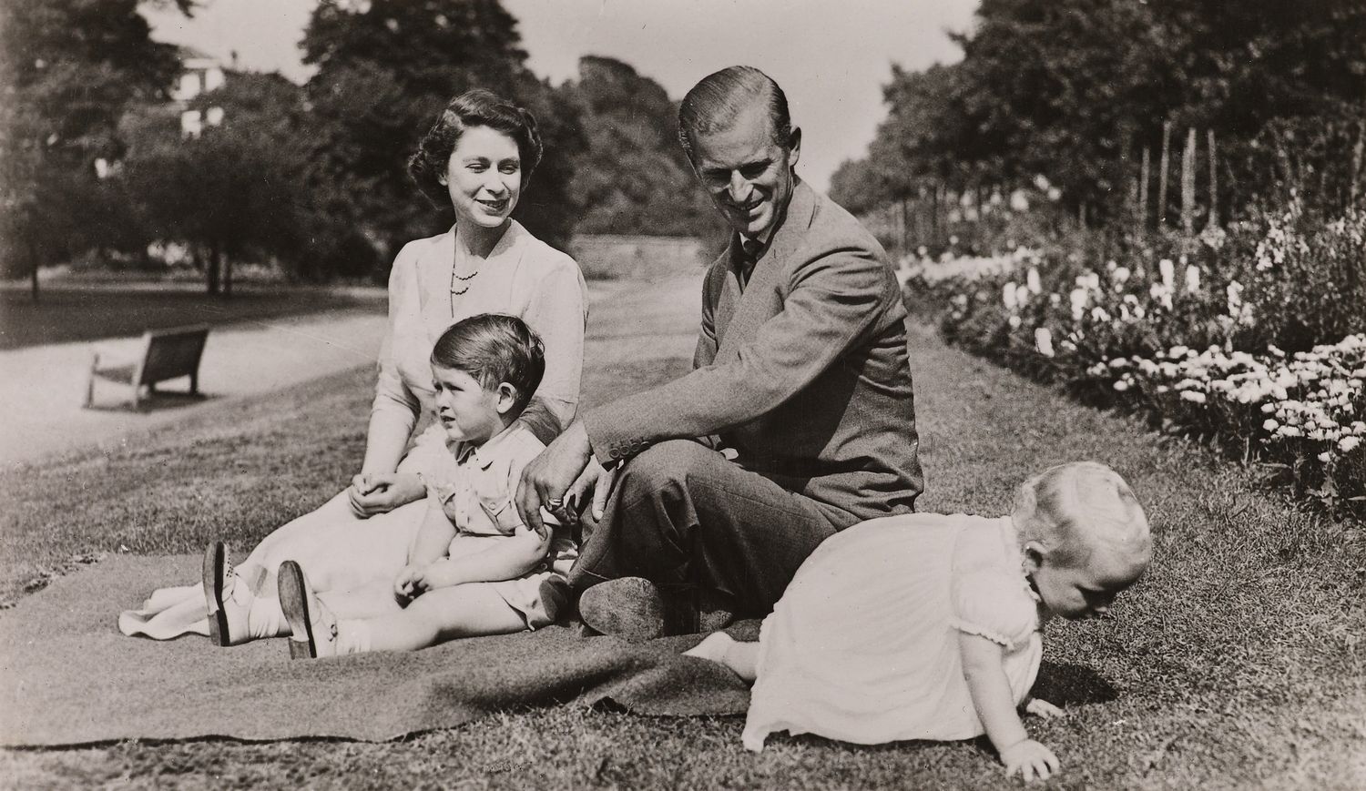 Photograph of HM The Queen (b. 1926) seated on the grass within the gardens of Clarence House. In front of her is seated HRH Prince Charles (b. 1948). HRH The Duke of Edinburgh (b. 1921) is seated on the Queen's left and looks at HRH Princess Anne (b. 195