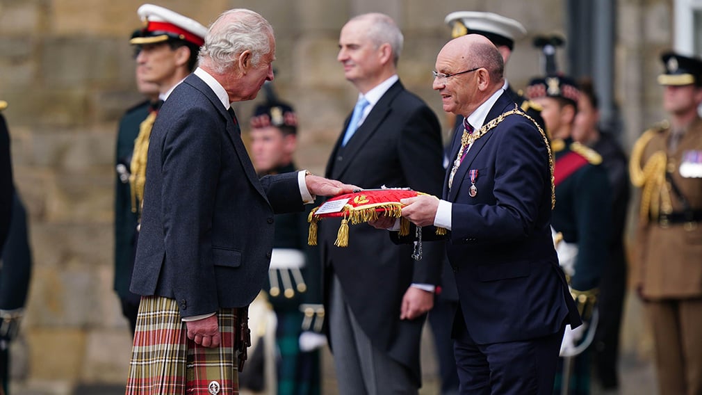 King Charles III receives the Keys to the City of Edinburgh from Lord Provost Councillor Robert Aldridge. King Charles III is wearing a kilt and is placing his hand on a red velvet cushion held out n front of him.