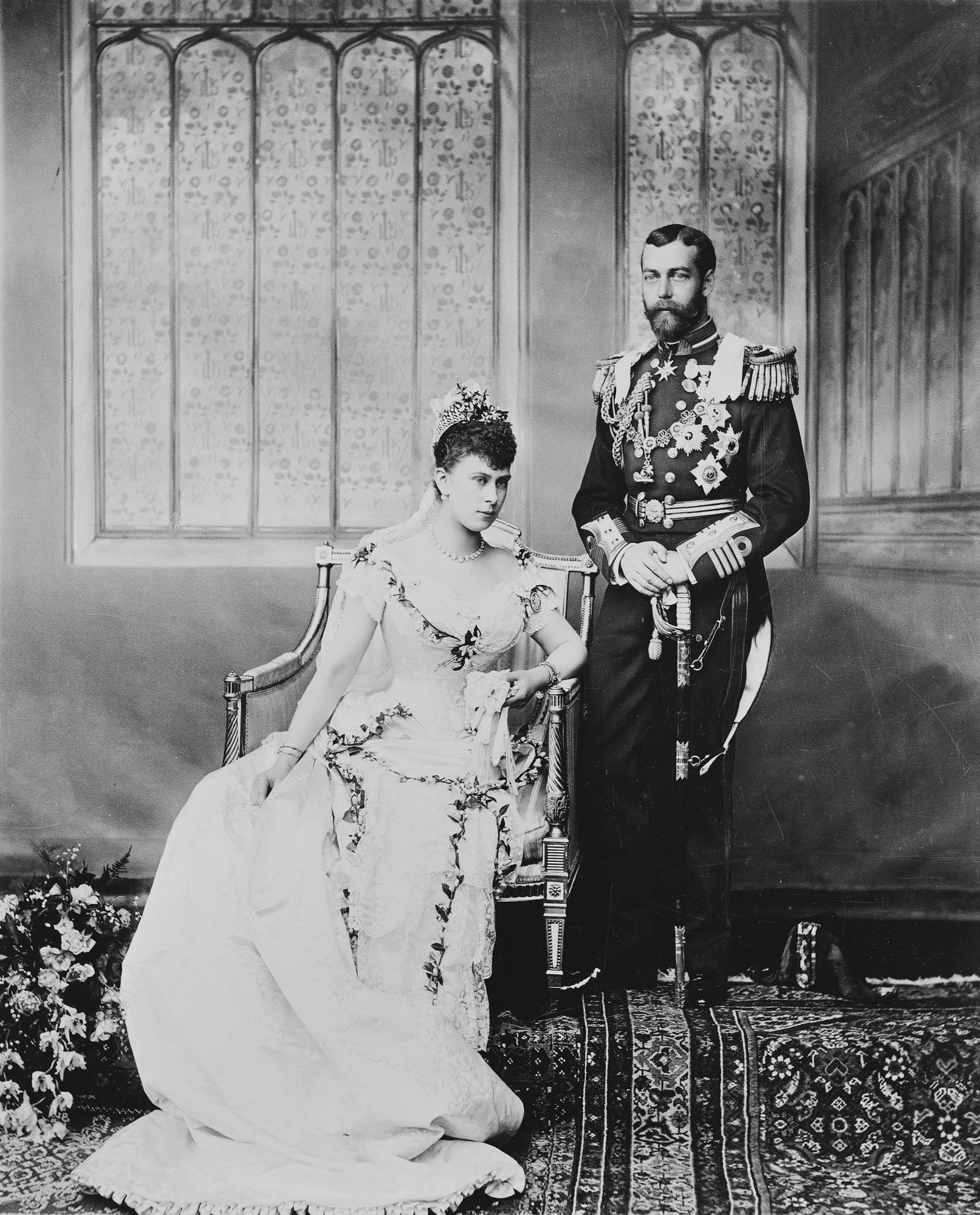 Photograph of the Duke and Duchess of York, later King George V (1865-1936) and Queen Mary (1867-1953): full length portrait, Princess Mary seated in her wedding gown of ivory silk satin, trimmed with Honiton lace and orange blossom. The Duke in full mili