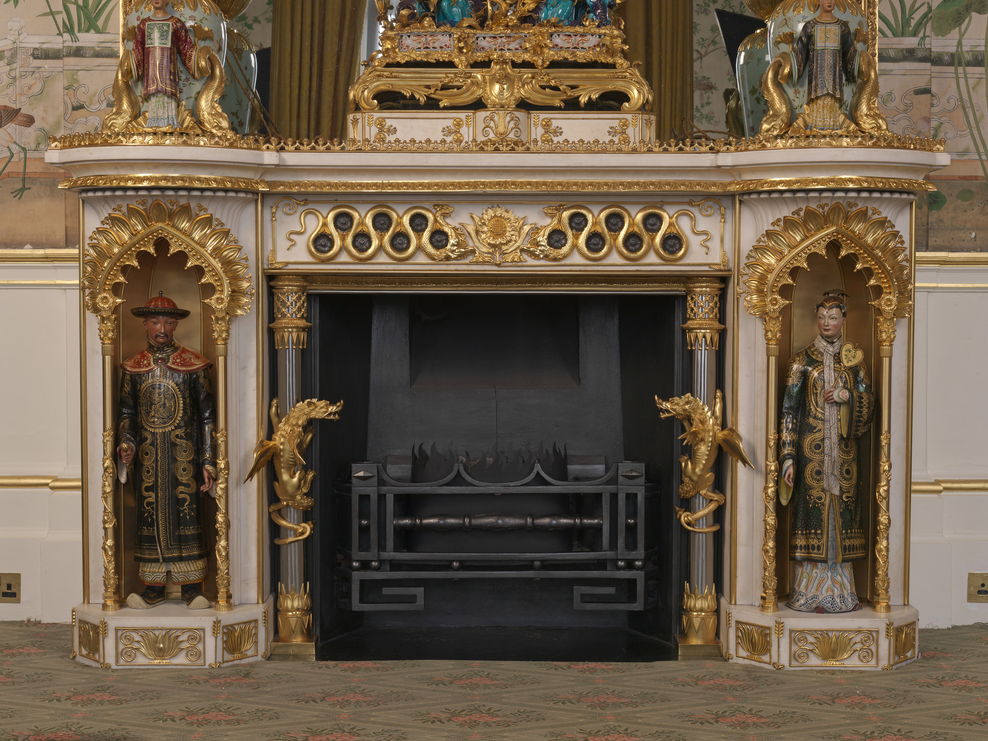 A white marble fireplace surround, with projecting rounded ends, above two&nbsp;japanned metal figures, in Chinese-inspired dress,&nbsp;of a man and a woman, standing left and right respectively in niches, with painted gilt-bronze arches edged with stylis