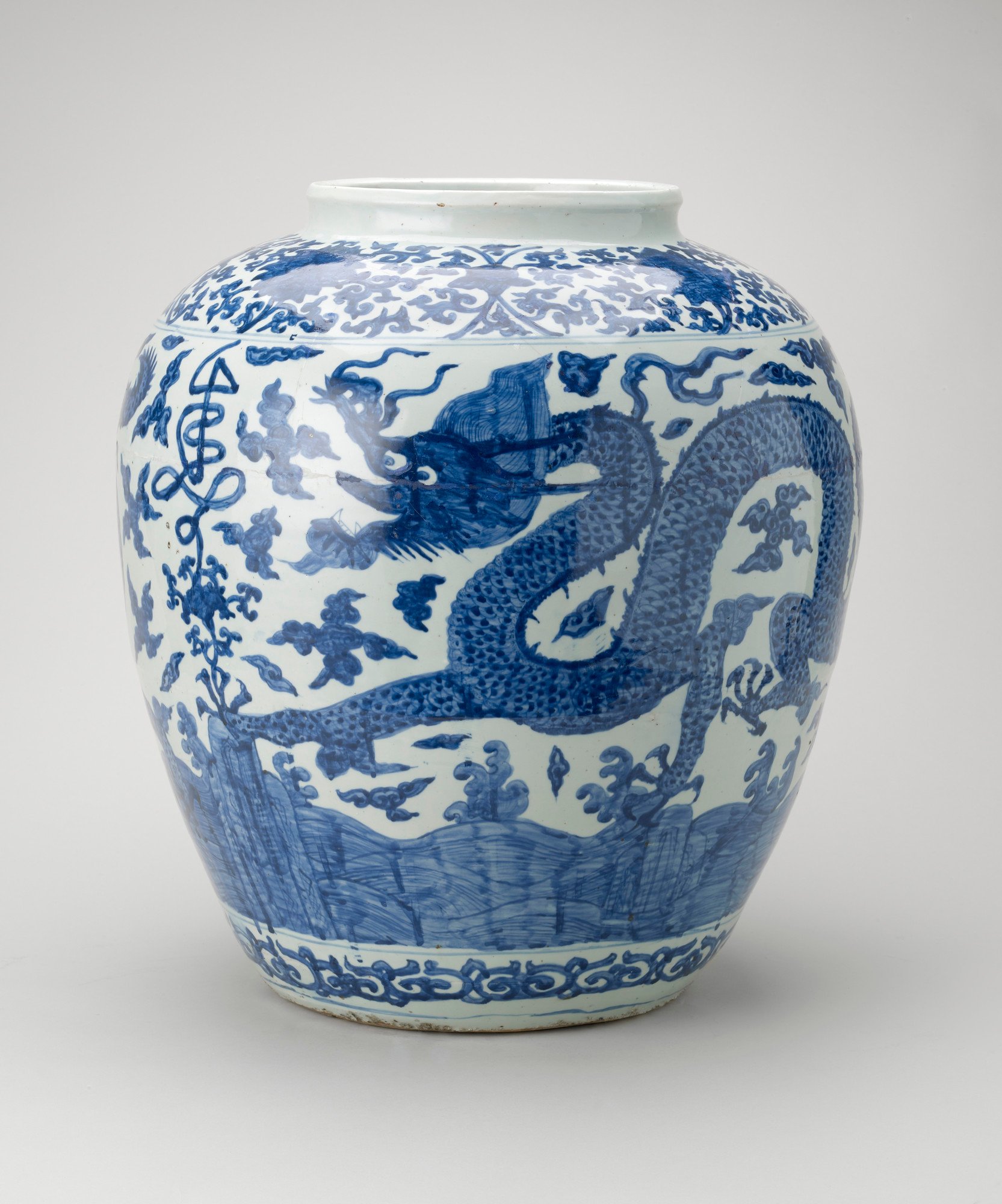 An ovoid-shaped Chinese Ming period porcelain jar painted in rich blue around the sides with two five-clawed dragons among clouds and with rocks and waves below. Round the shoulder a stylised shou (long life character) seems to grow out of the lotus scrol