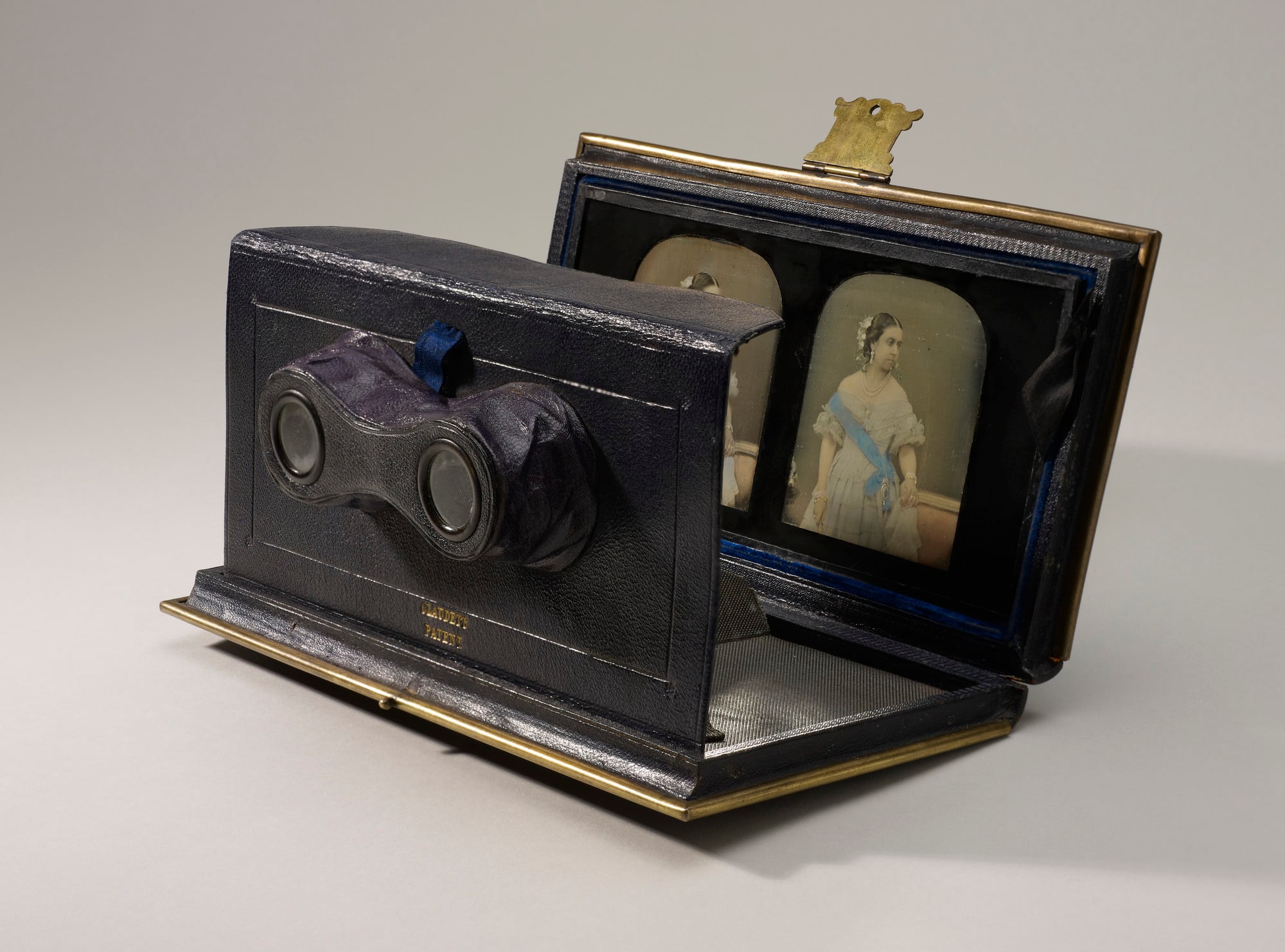 Hand-tinted stereoscopic daguerreotype of Queen Victoria. The image shows her standing with her head turned to the right and her left hand resting on the chair beside her. She is wearing a pale blue dress with a deeper blue sash and has flowers in her hai
