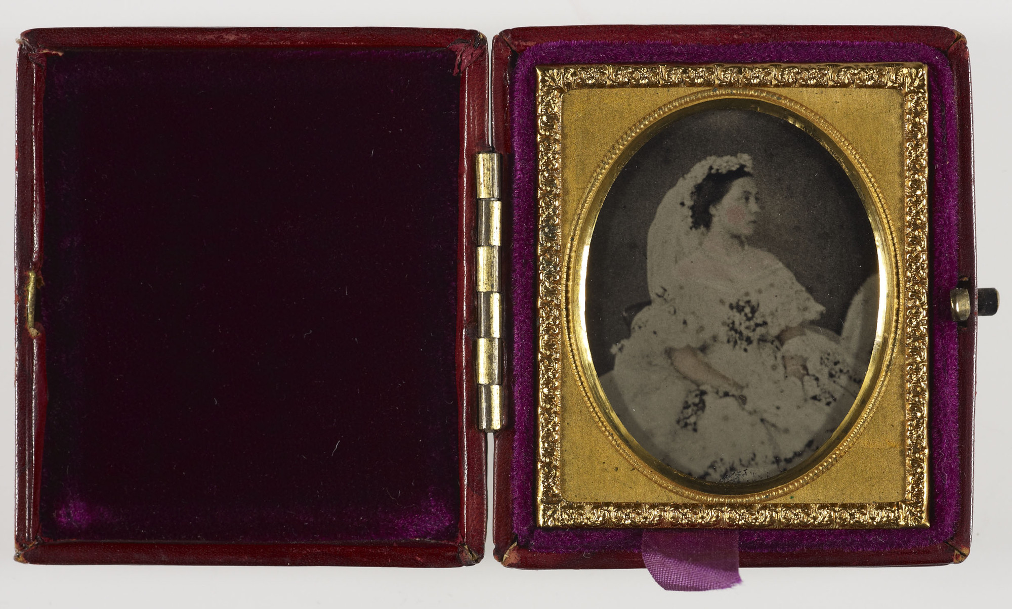Hand-tinted ambrotype of Princess Victoria wearing her wedding dress. She is seated facing right wearing a white lace dress decorated with flowers and a veil. The ambrotype is mounted in a gilt frame housed in a brown leather case with a red velvet interi