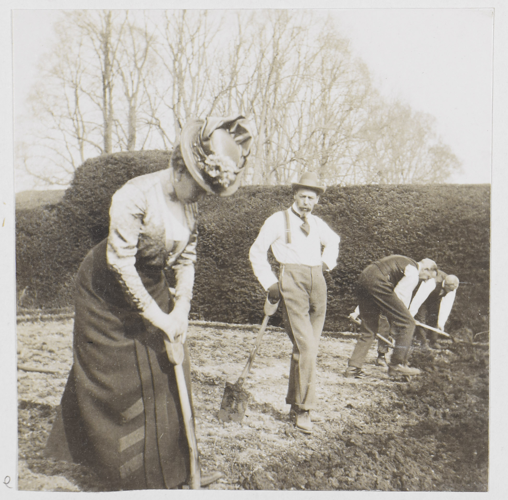 Photograph of Queen Mary (1867-1953),standing closest to the viewer whilst she digs with a spade within a potato plot in Windsor, probably the Home Park.&nbsp;On her left stands King George V (1865-1936), wearing shirt sleeves and a homburg hat, leaning o
