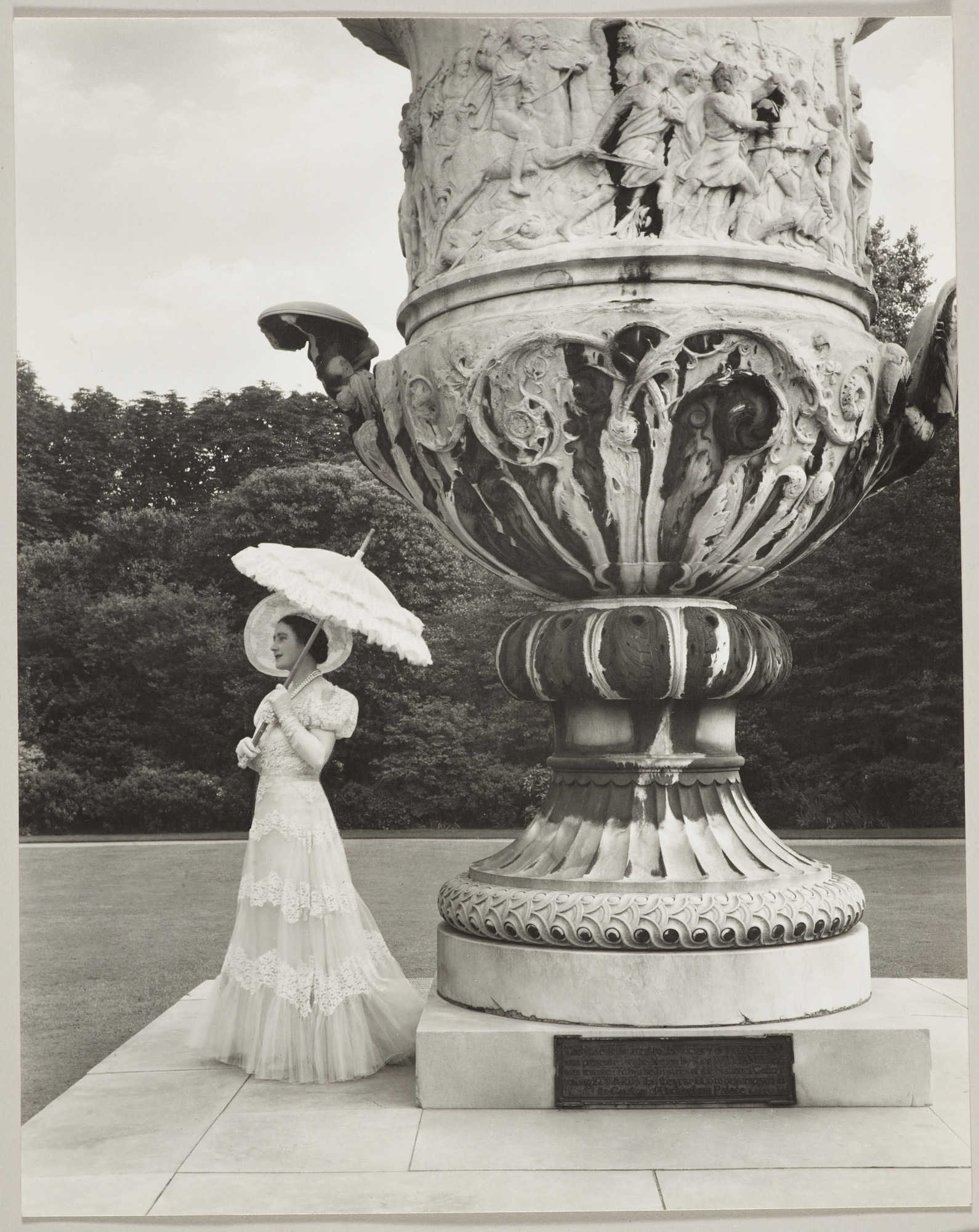 'Queen Elizabeth, Buckingham Palace Gardens' (RCIN 2315149) by Cecil Beaton Photograph of Queen Elizabeth, later Queen Elizabeth The Queen Mother (1900-2002) standing beside the Waterloo Vase on the grounds of Buckingham Palace. She faces three quarters to the left and holds a parasol. She wears lon