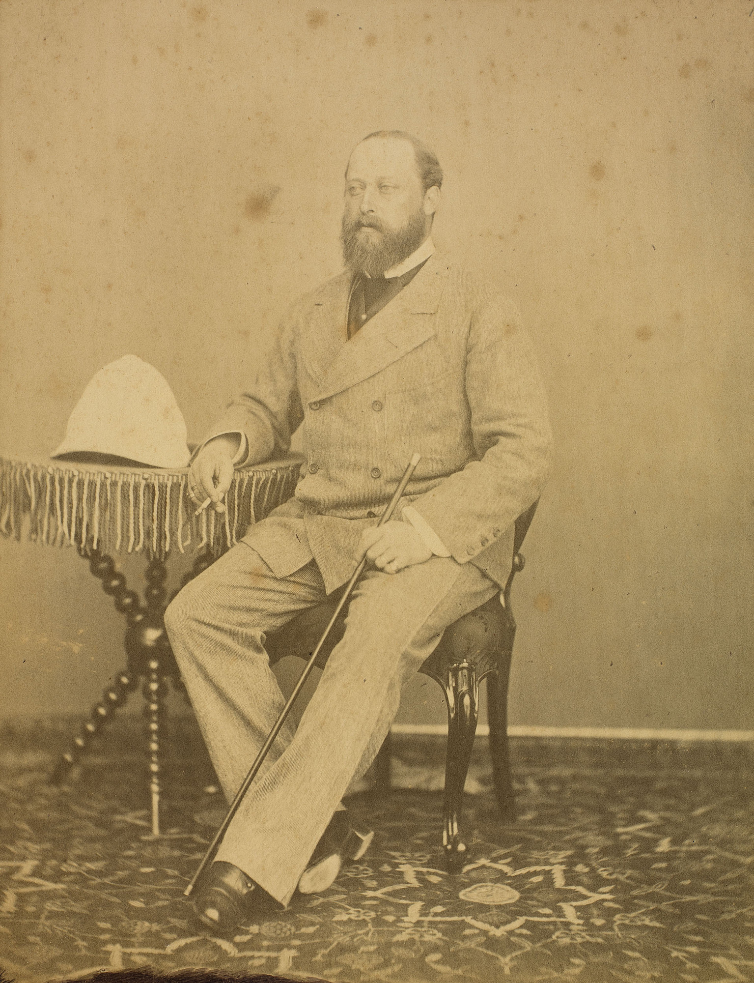 Photograph showing full length portrait of Albert Edward, Prince of Wales, seated, holding a cane in his left hand and a cigarette in his right, leaning on a small, three-legged table on which is a pith helmet. The Prince is wearing trousers with a pronou