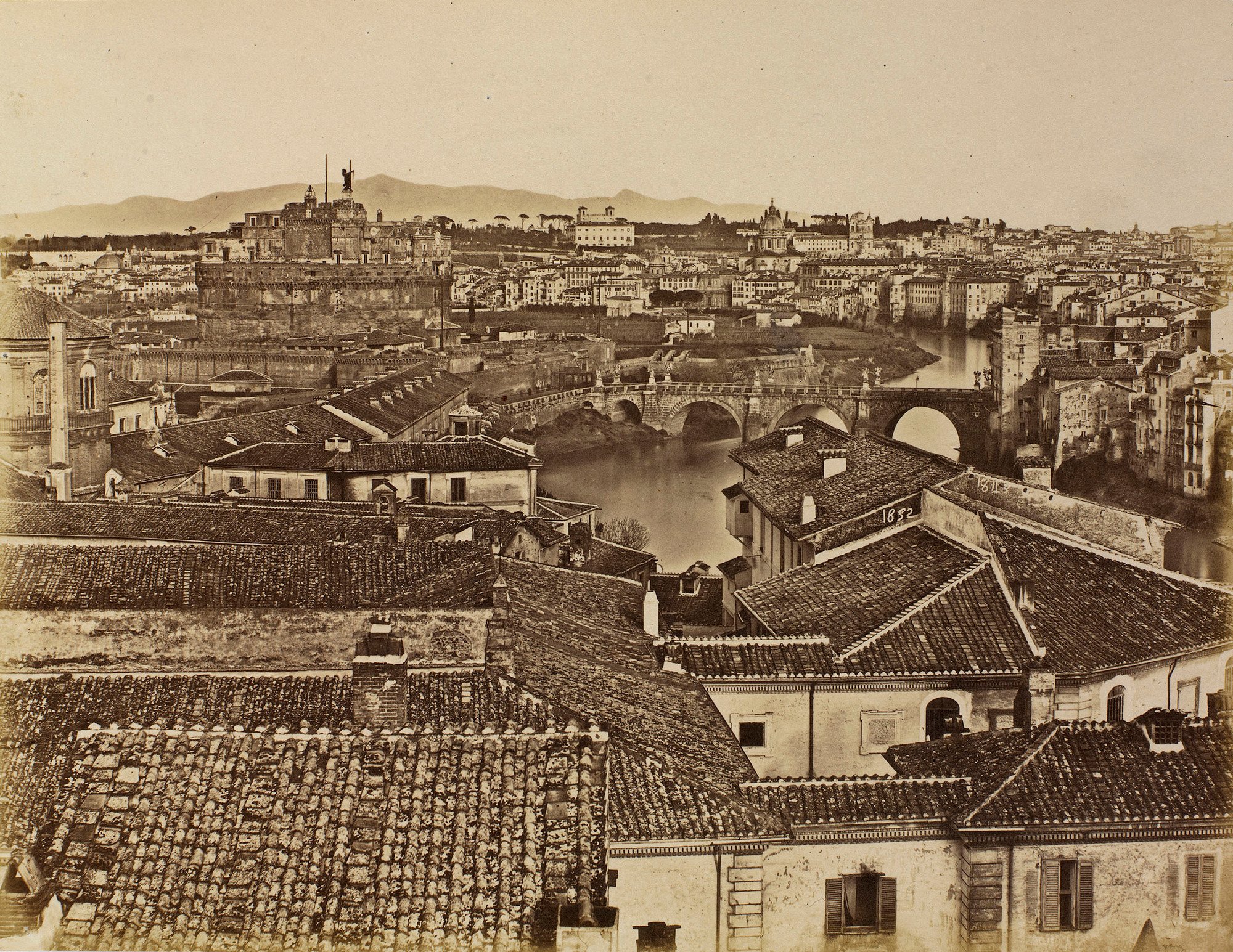 Photograph of a view of Rome with the bridge of St Angelo spanning the river Tiber in the middle distance and the Sabine Hills in the far distance. Roof tops of buildings dominate the foreground.