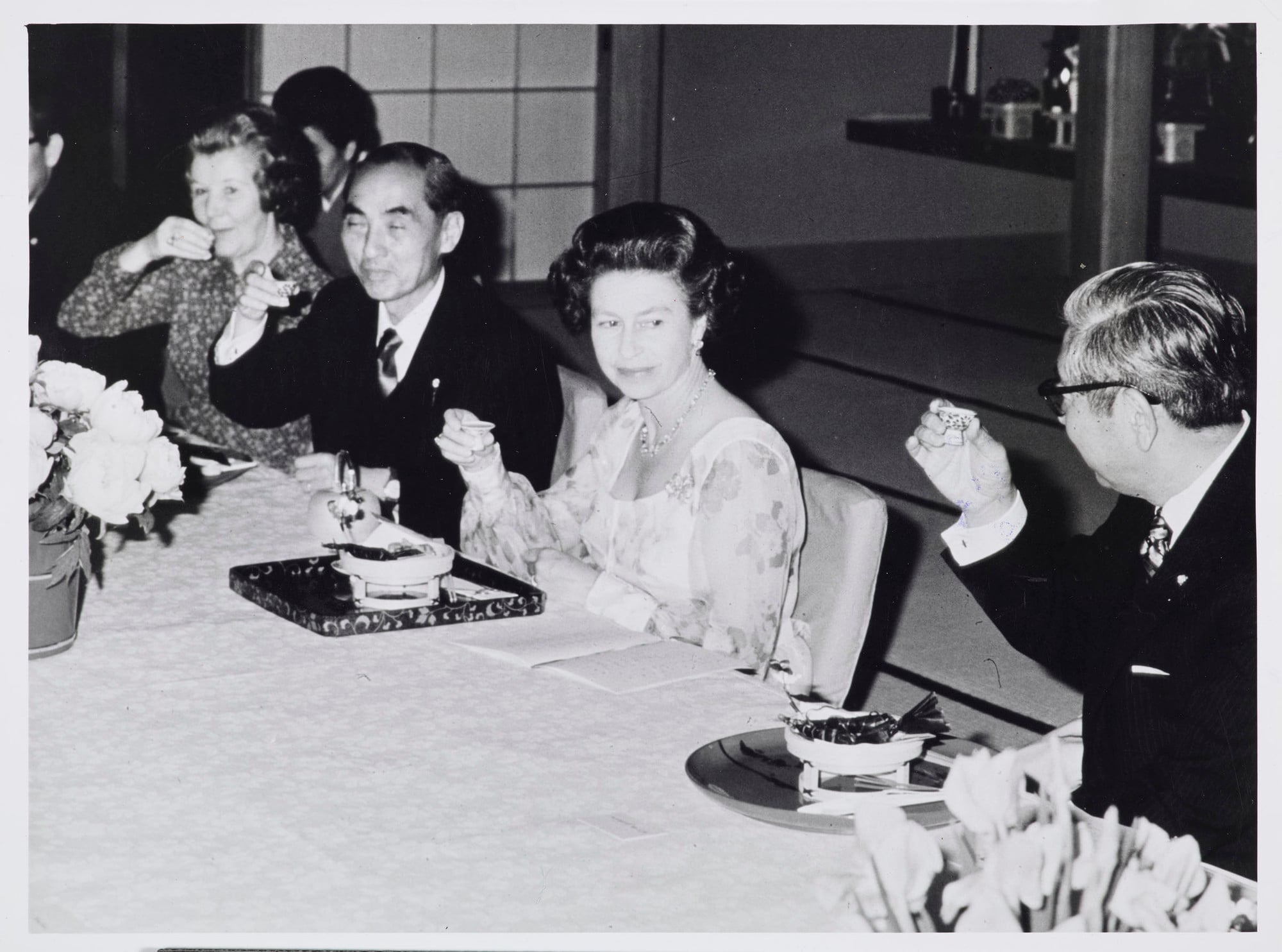 Queen Elizabeth II drinking Sake during dinner at a Japanese restaurant in Kyoto Photograph of Queen Elizabeth II sitting on a Japanese-style on a tatami floor mat whilst drinking saké at a restaurant in Kyoto. The photograph was taken during the Queen's six-day State Visit to Japan in 1975. Lady Shepherd and Mr