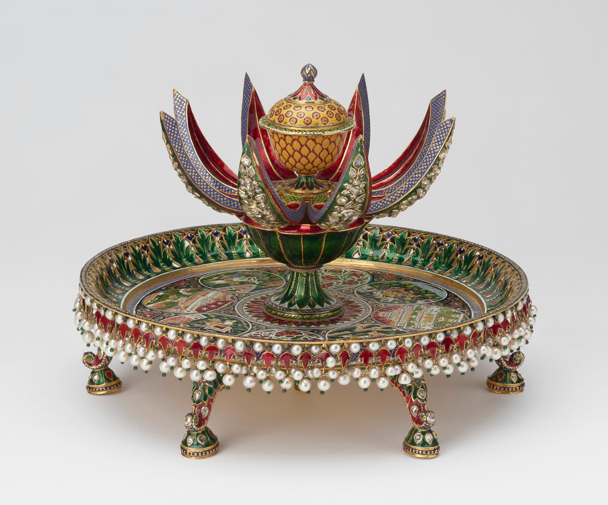 Perfume holder An enamelled gold perfume holder or attardan formed of a circular tray with a green and red enamelled gold lotus flower in the centre that opens to reveal a yellow and red cup and cover when pressure is applied to the rod beneath the tray. The tray is dec