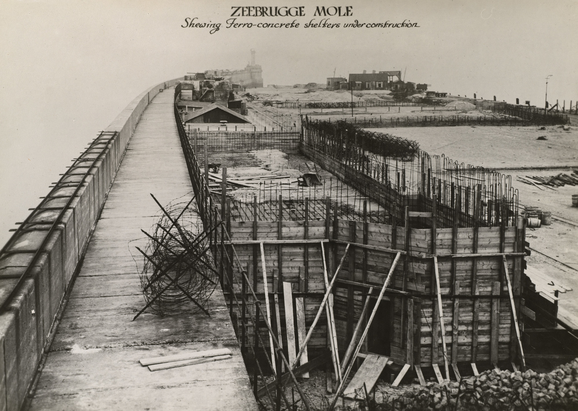 Zeebrugge Mole Photograph of a view along a harbour wall, the Zeebrugge Mole, Belgium. On the right are a series of ferro-cement shelter under construction. A mass of barbed wire sits on the harbour wall. A light house is positioned at the end of the harbour w