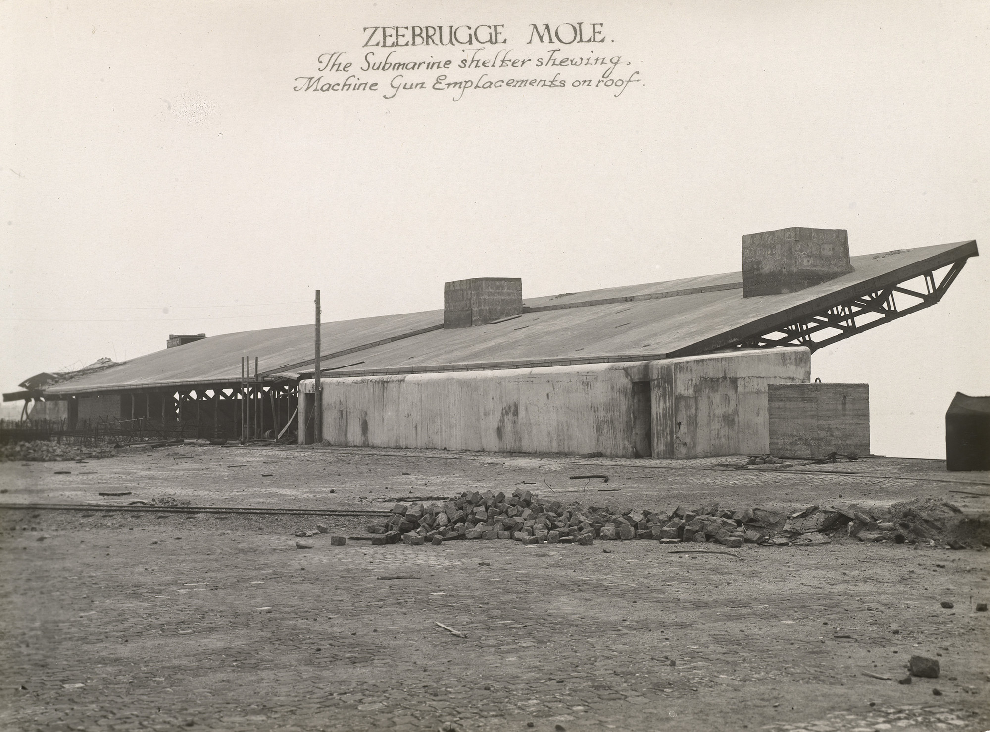 Photograph of a view over a cobbled yard,&nbsp;leading towards a concrete submarine shelter featuring machine gun emplacements on roof