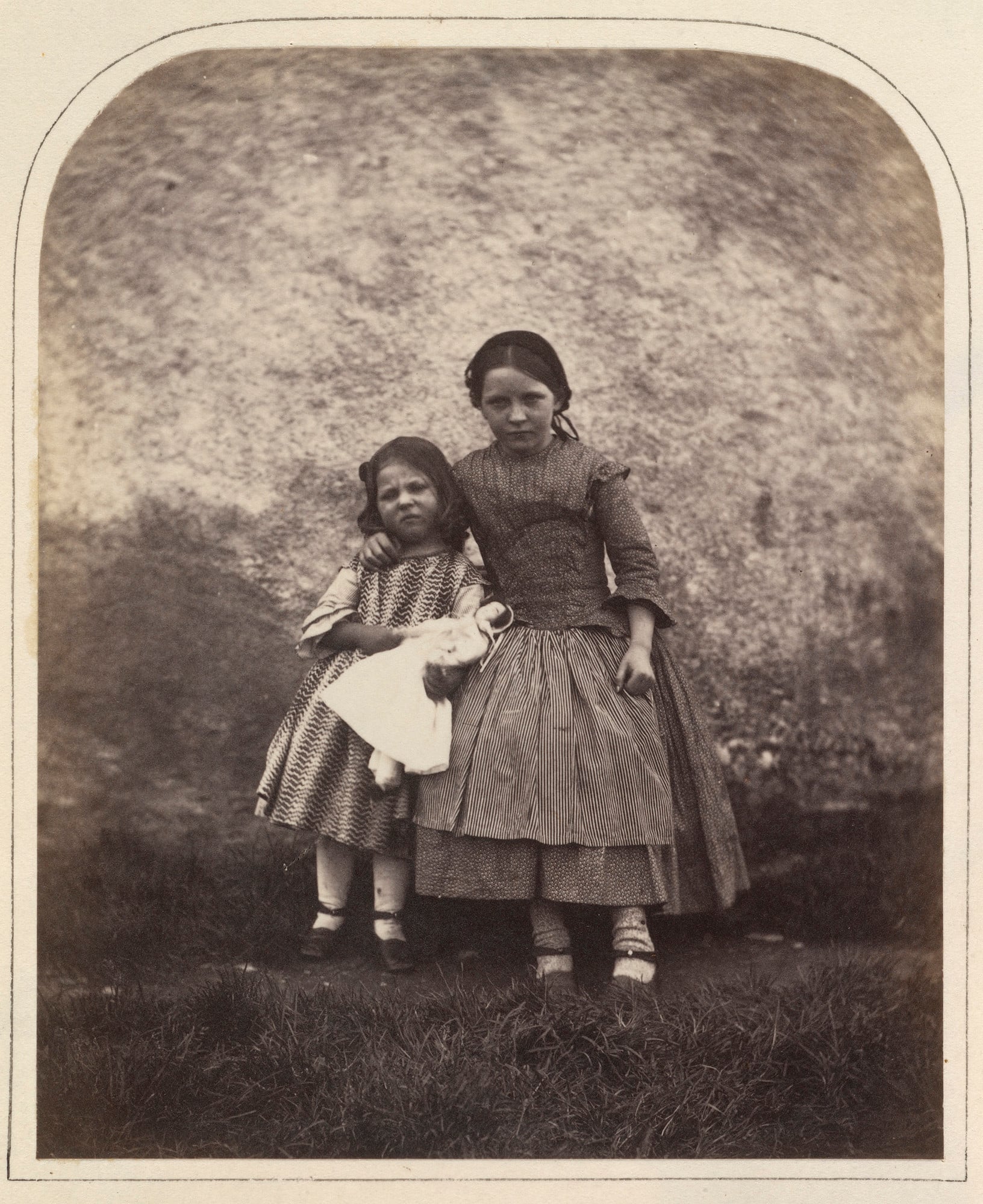 Photograph of a full length double portrait of the sisters, Victoria-Alice and Mary Symons. The young girls stand in front of a wall and face the camera front on. The younger girl holds a doll. They both wear patterned dresses, light colour stockings and 