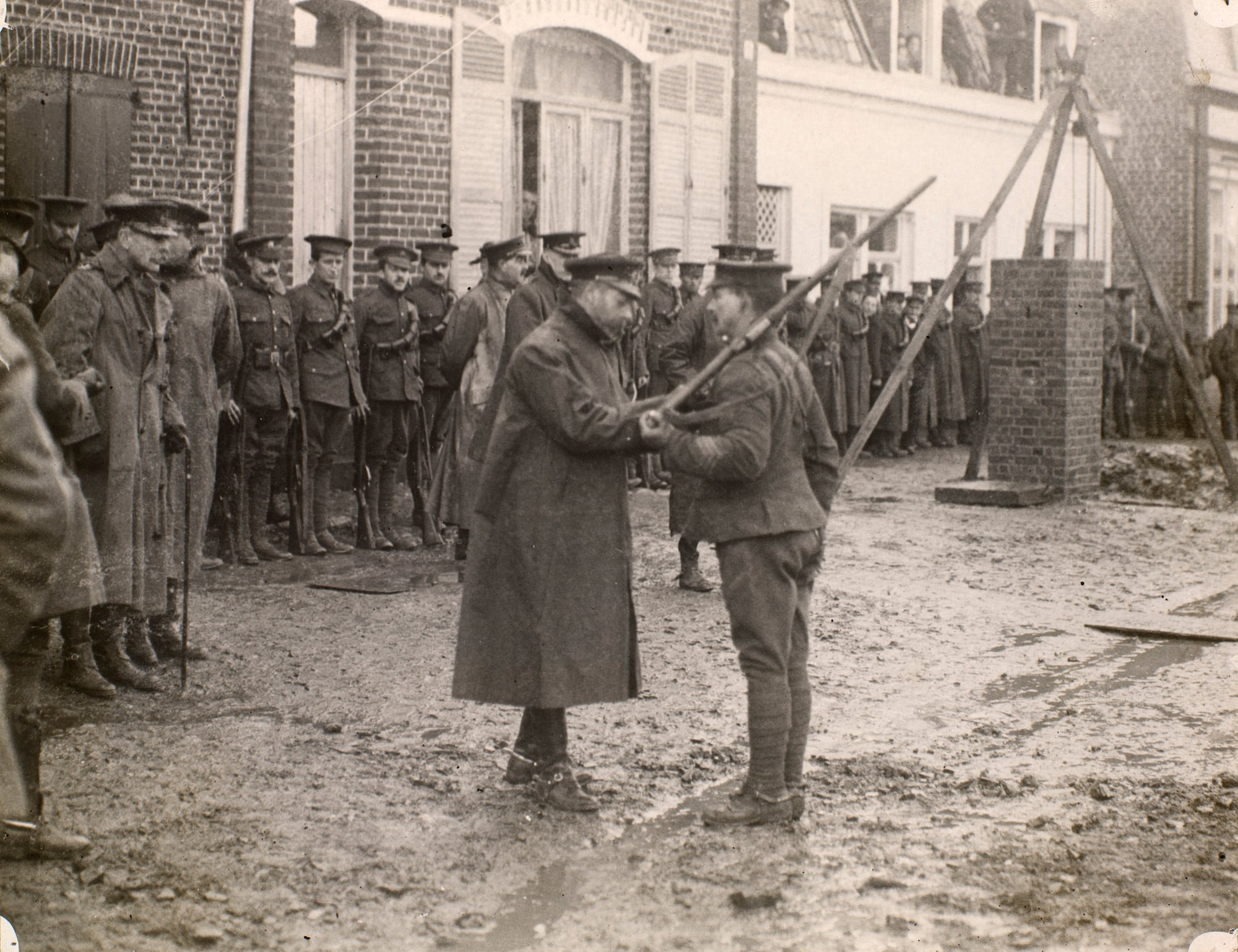 King George V (1865-1936) distributing decorations to the 1st Division Photograph of King George V (1865-1936) in uniform and coat pinning a medal to the chest of a Sergeant from the 1st Division. The Sergeant holds a rifle over his left shoulder. The action takes place on a muddy street lined with buildings. A lin