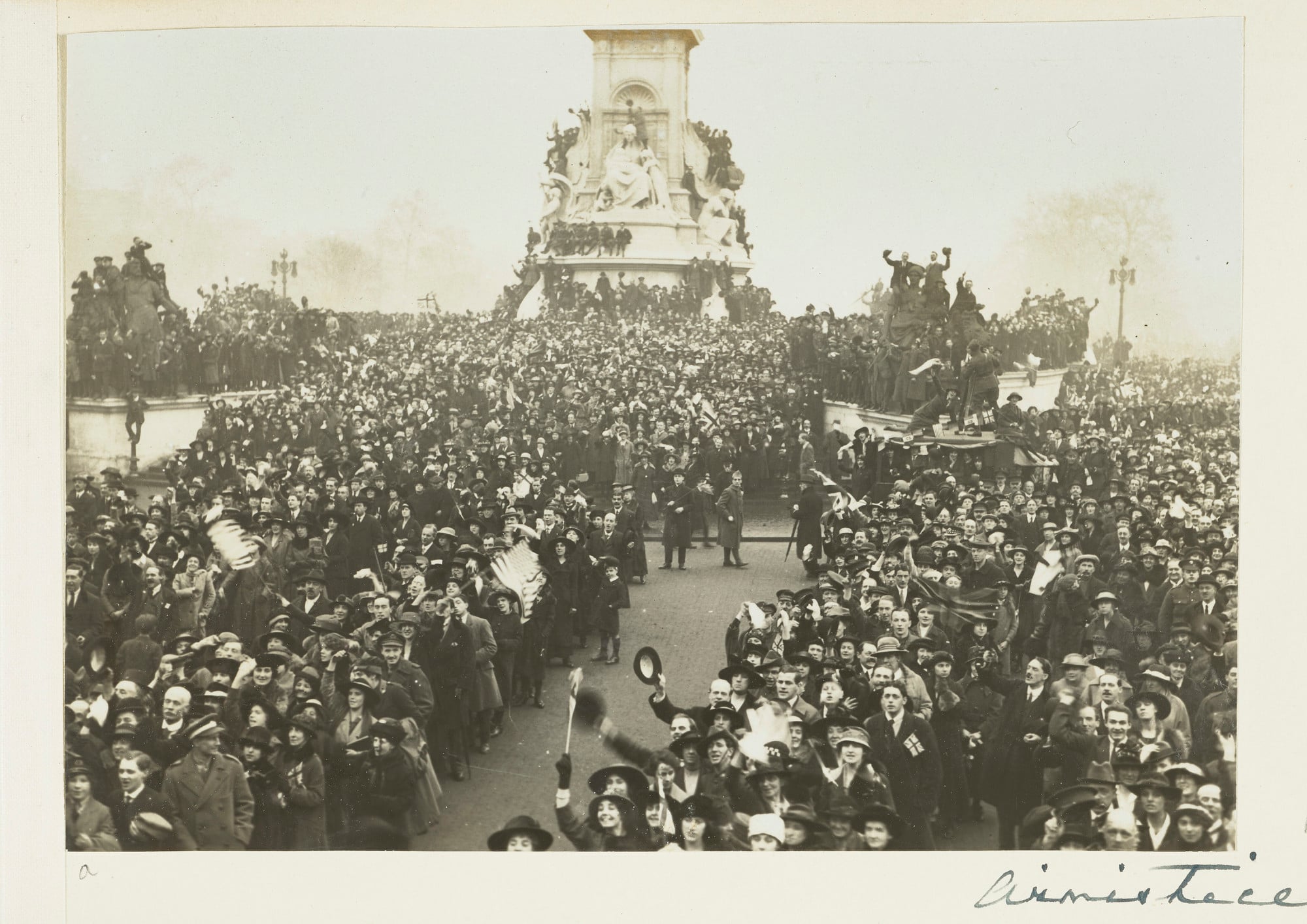 Photograph of a large crowd gathered outside of Buckingham Palace to celebrate the announcement of the end of World War I. The crowd stand around and sit on the Queen Victoria monument. Soldiers and civilians are shown cheering and waving British and Amer
