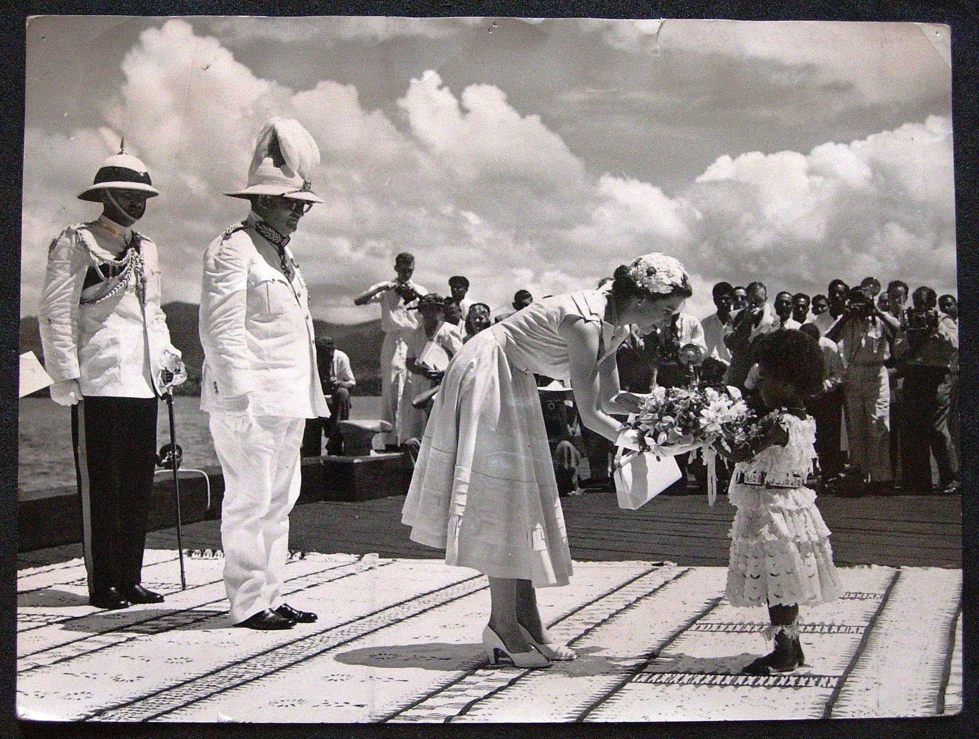 Adi Kainona presents The Queen with a bouquet of Fijian flowers Photograph showing a Fijian child, Adi Kainona, presenting The Queen with a bouquet of Fijian flowers on The Queen's arrival at Suva Wharf from the SS Gothic. Behind the Queen stands Sir Ronald Garvey, Governor of Fiji and another man in uniform. Pho