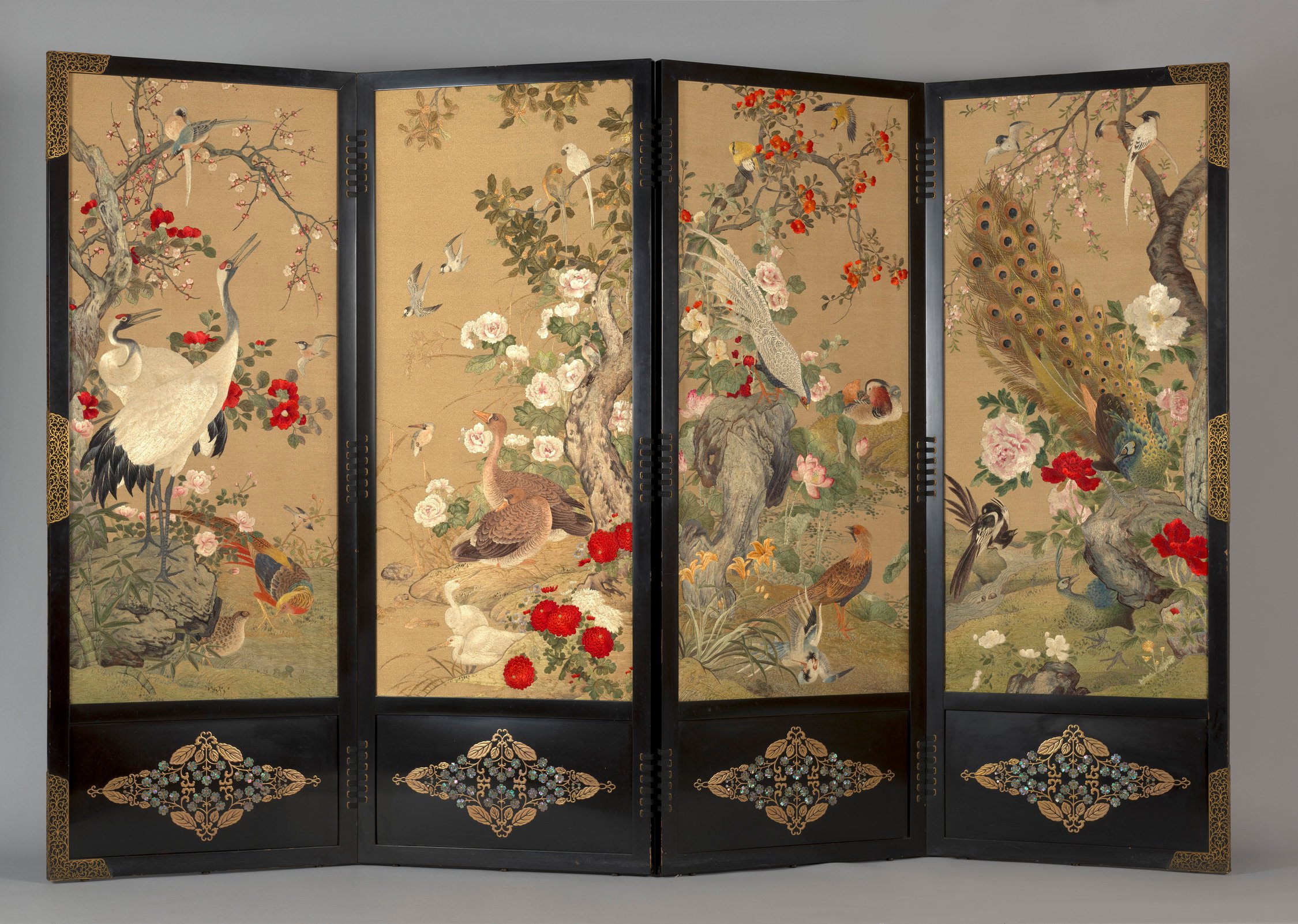 Each of the four panels is fitted with cream silk, which is richly embroidered. The first panel features a pair of cranes standing on rocks and grass, with a pheasant underneath tree peony and cherry-blossom branches. The glossy feathers of the cranes are