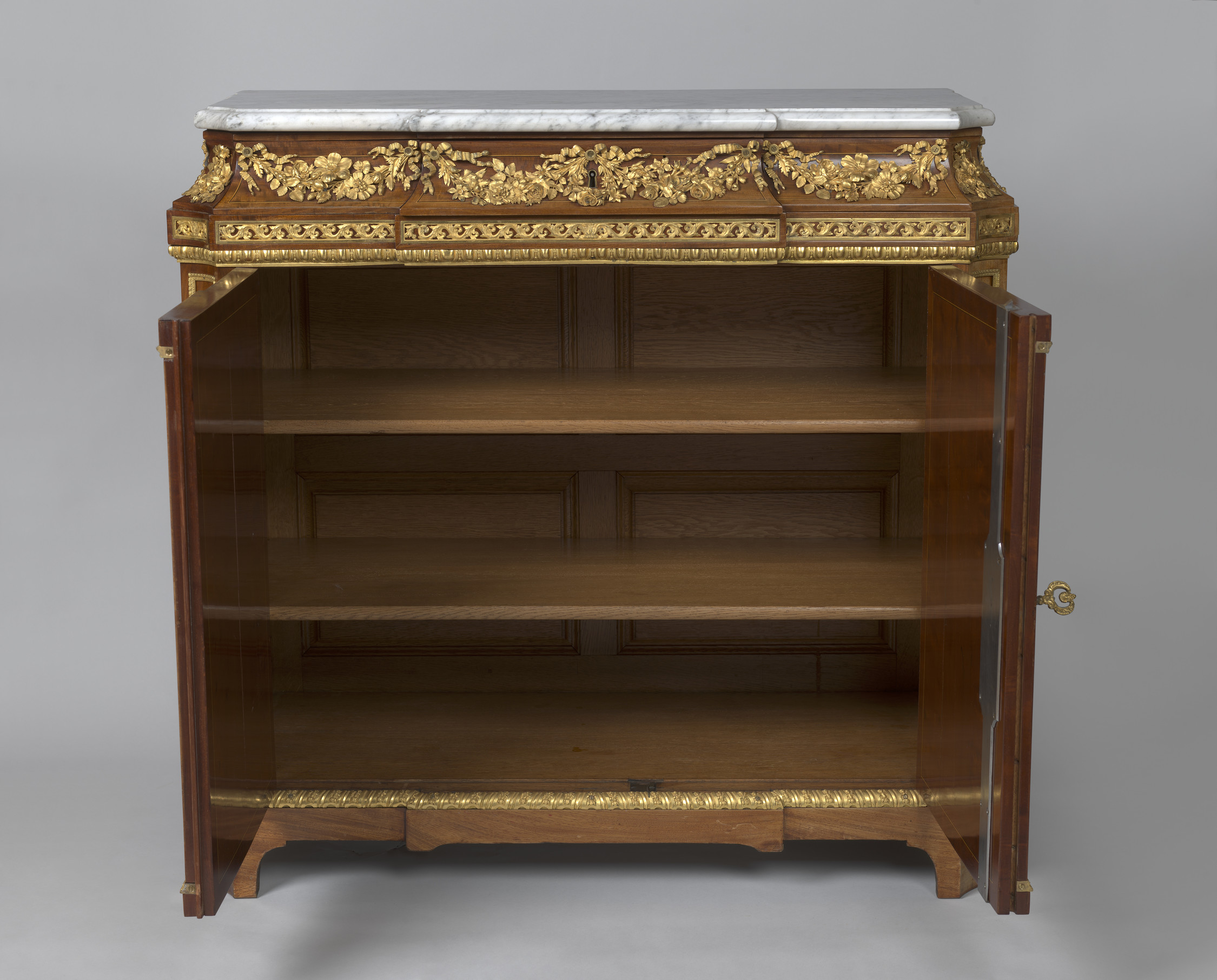 Rectangular cabinet veneered in mahogany with white marble top and gilt bronze mounts. The concave frieze above two doors with gilt bronze mounts of garlands of flowers, the door&nbsp;panels edged with gilt&nbsp;bronze foliage mounts. The angles with cant