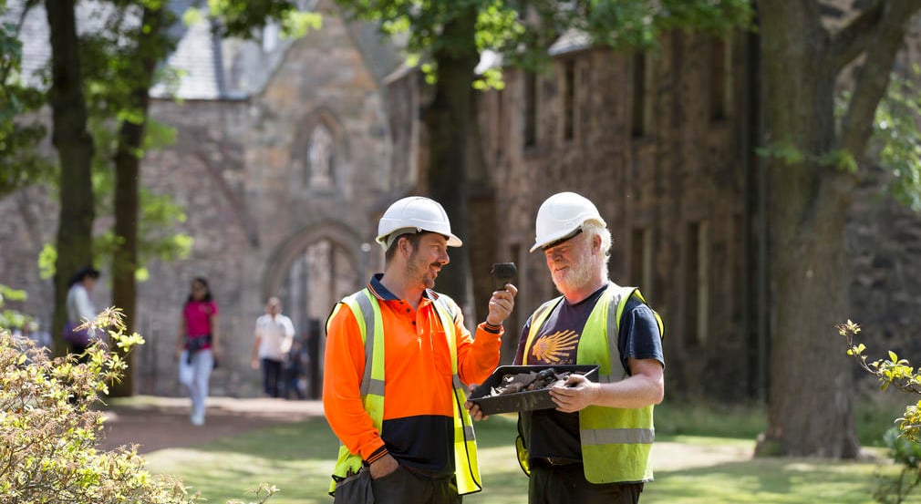 Archeologists at the Palace of Holyroodhouse. Two archeologists in high-vis vests stand outside the Palace of Holyroodhouse while looking at finds in a tray.