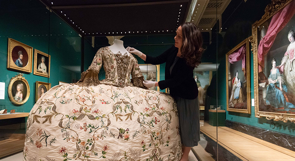 Georgian dress on display. A wide hooped dress in a display cabinet in the exhibition. A curator adjusts the dress.