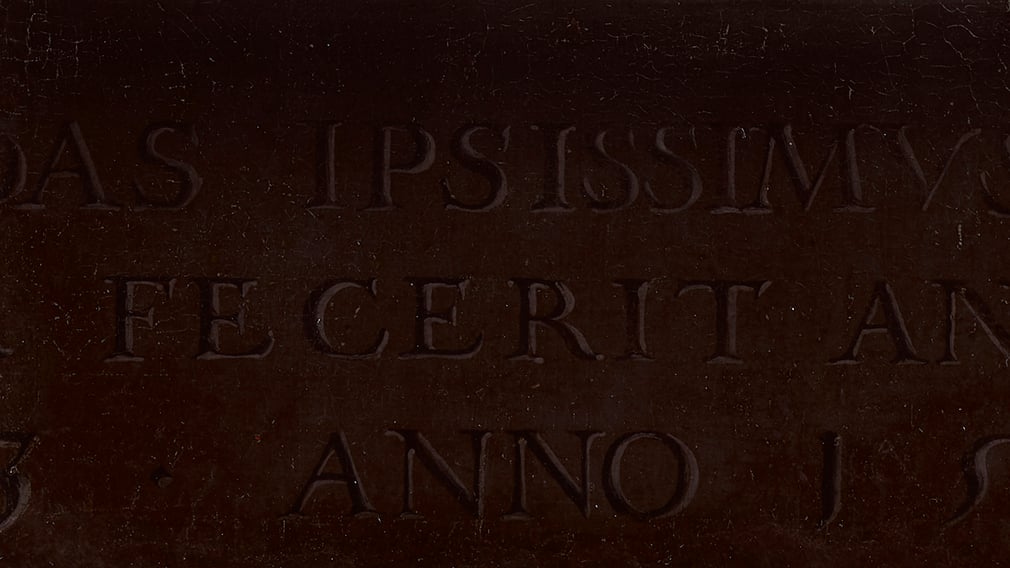 Detail of the 'carved' inscription. A latin inscription painted on a painting. The background is brown.
