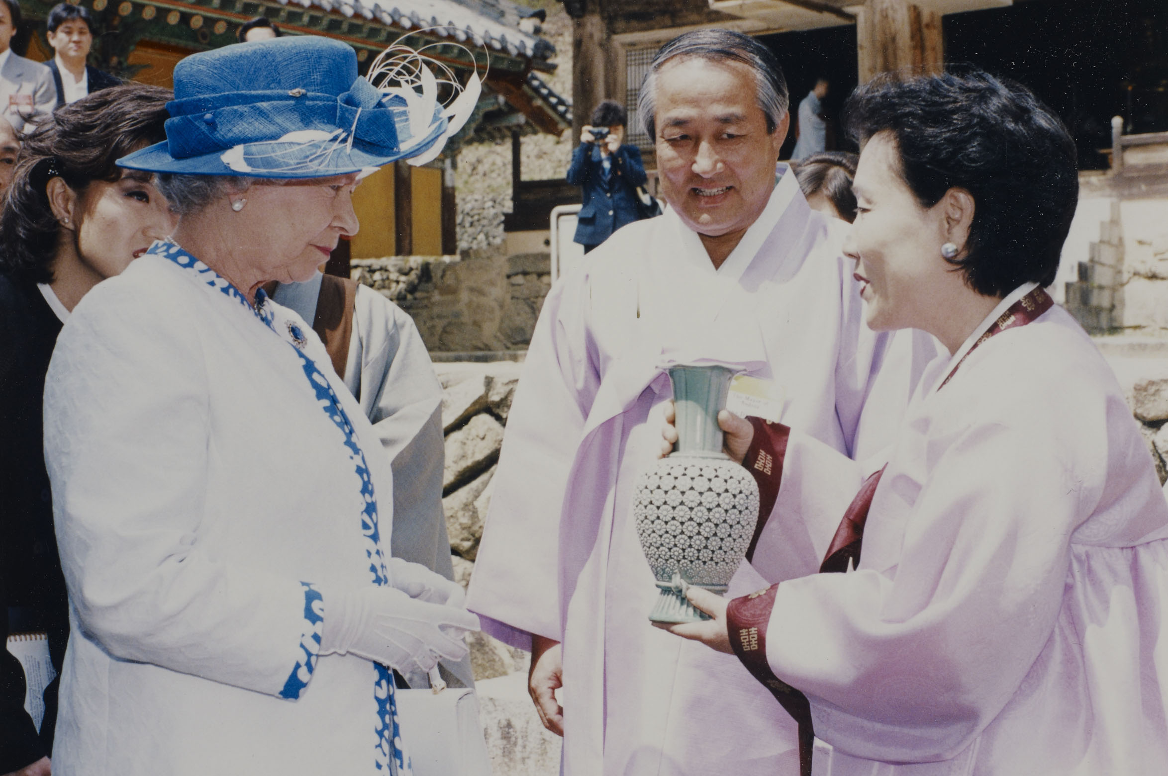 Photograph of Queen Elizabeth II receiving gift of a vase of green-grey celadon from the citizens and Mayor of Andong (RCIN 2361273). Photograph of Queen Elizabeth II wearing blue and white outfit. There are two people wearing traditional Korean dress. One of these people hold a vase.