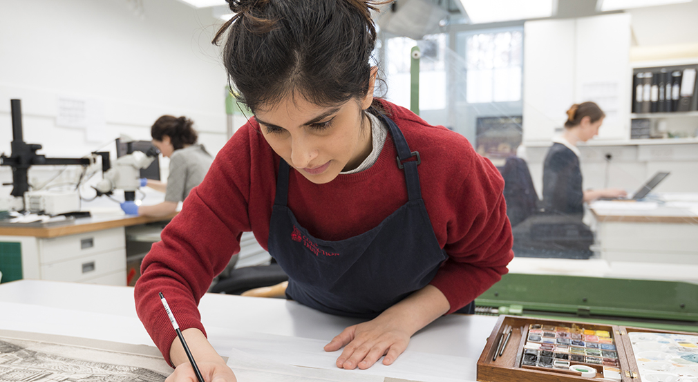 A person using a paintbrush to conserve an artwork. She is wearing red and has her hair up. 