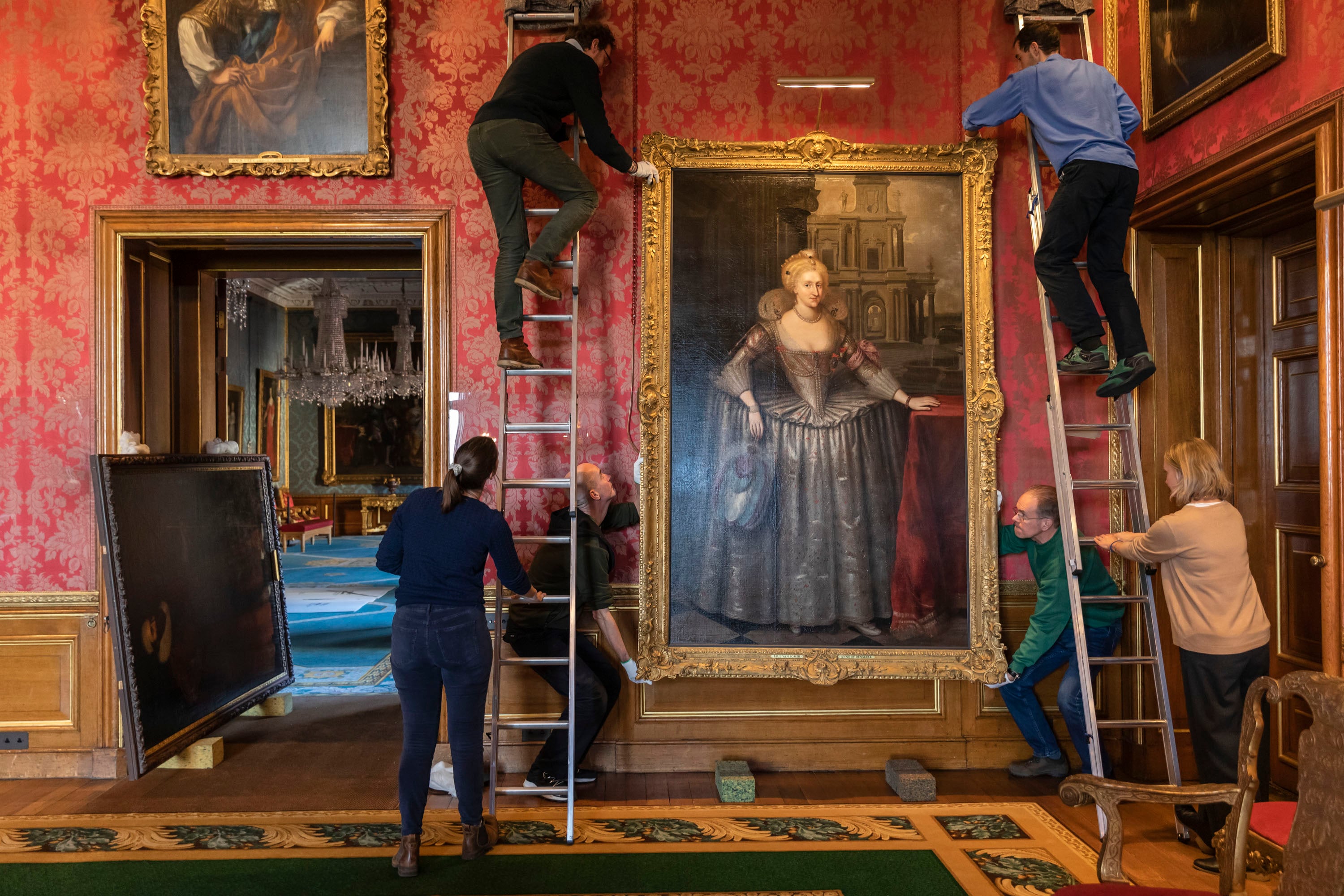 People position a large painting onto the wall. Two people are on ladders holding the painting, two people hold the painting at the bottom and two people are holding the ladders.