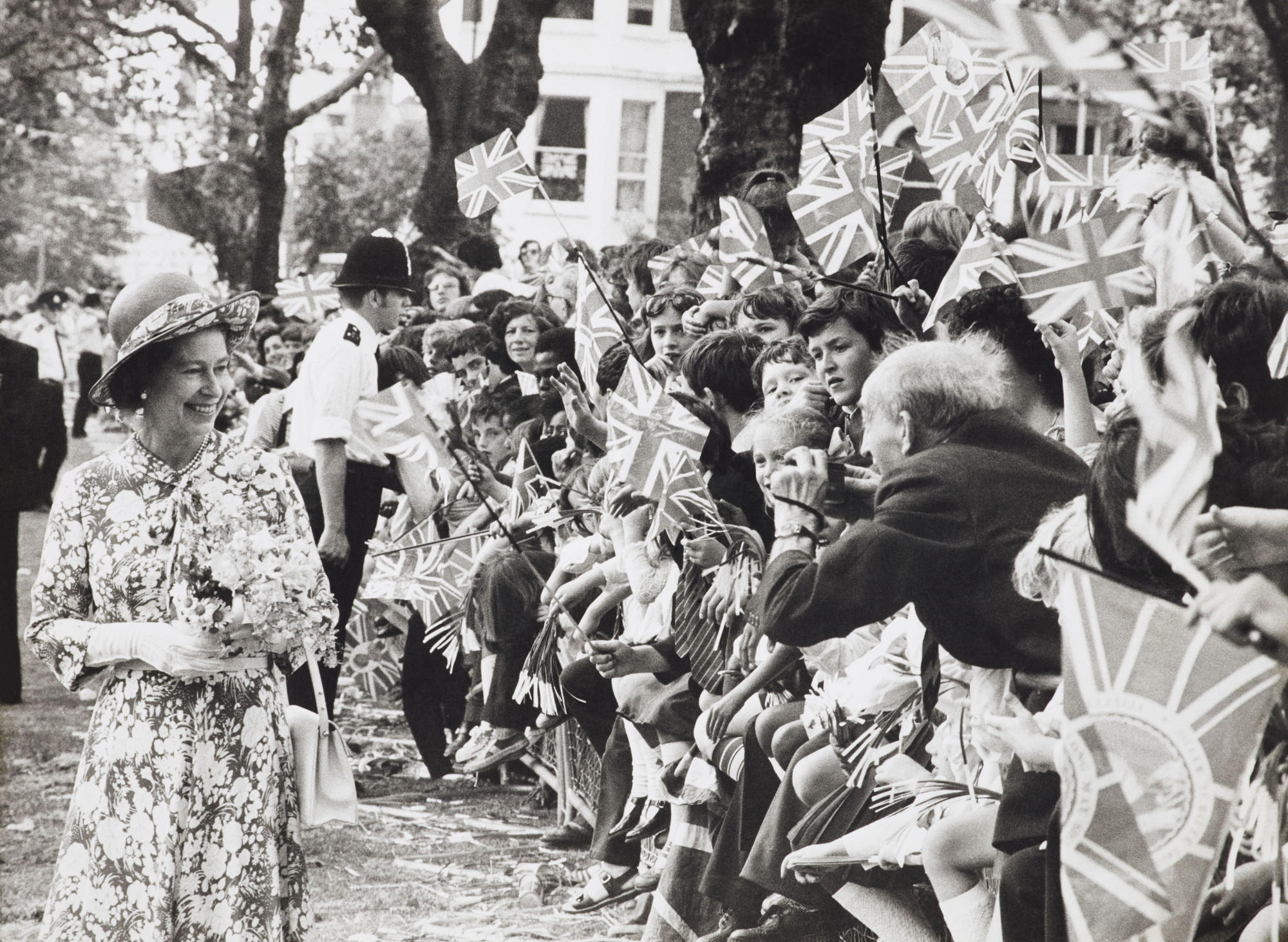 Photograph of HM Queen Elizabeth II (b.1926) visiting Highbury Fields during her Silver Jubilee tour of North London. She is walking to the left wearing a floral dress and holding a bouquet. There is a crowd of well wishers waving Union Jacks to the right
