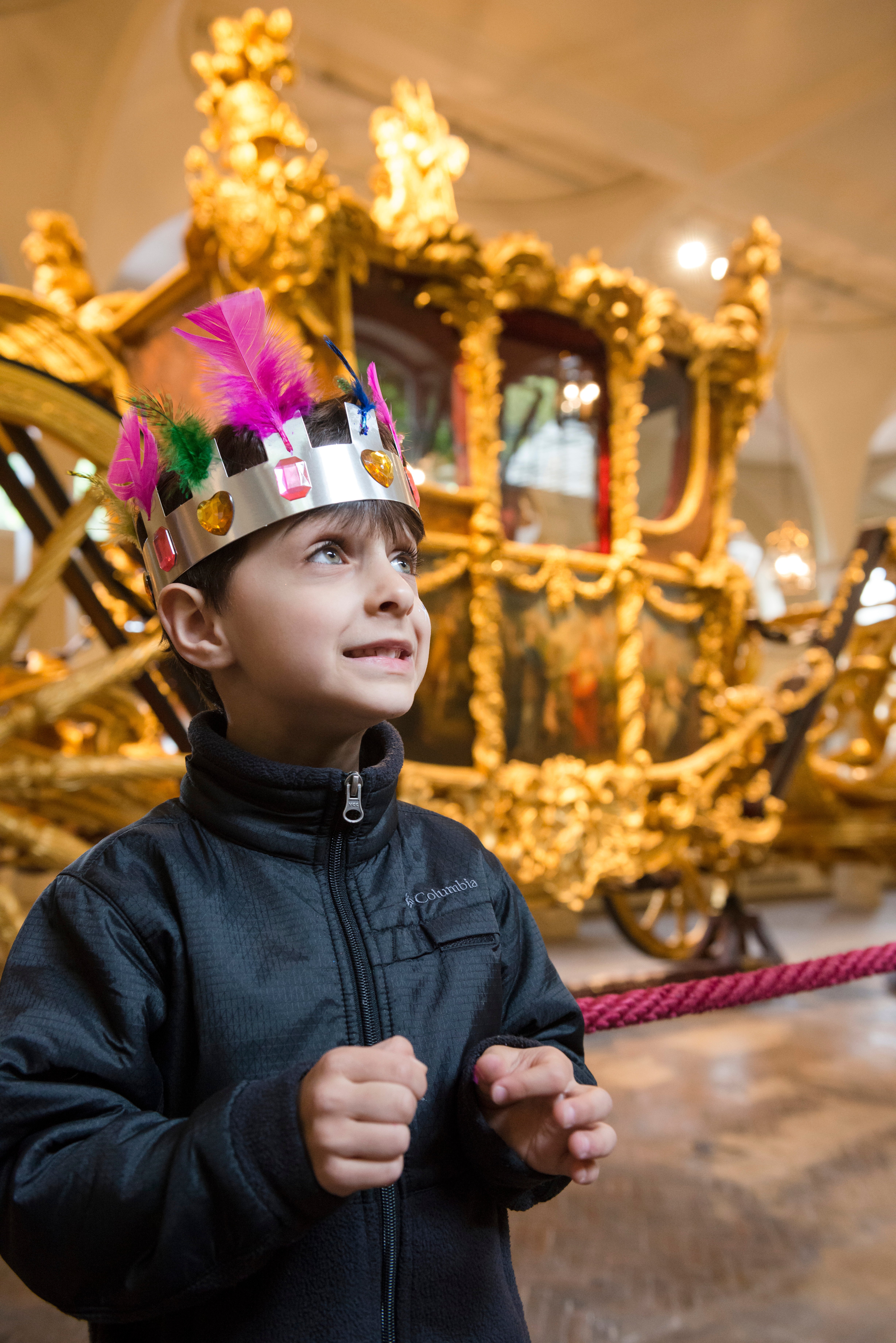 A child wearing a crown stands excitedly in front of the Gold State Coach