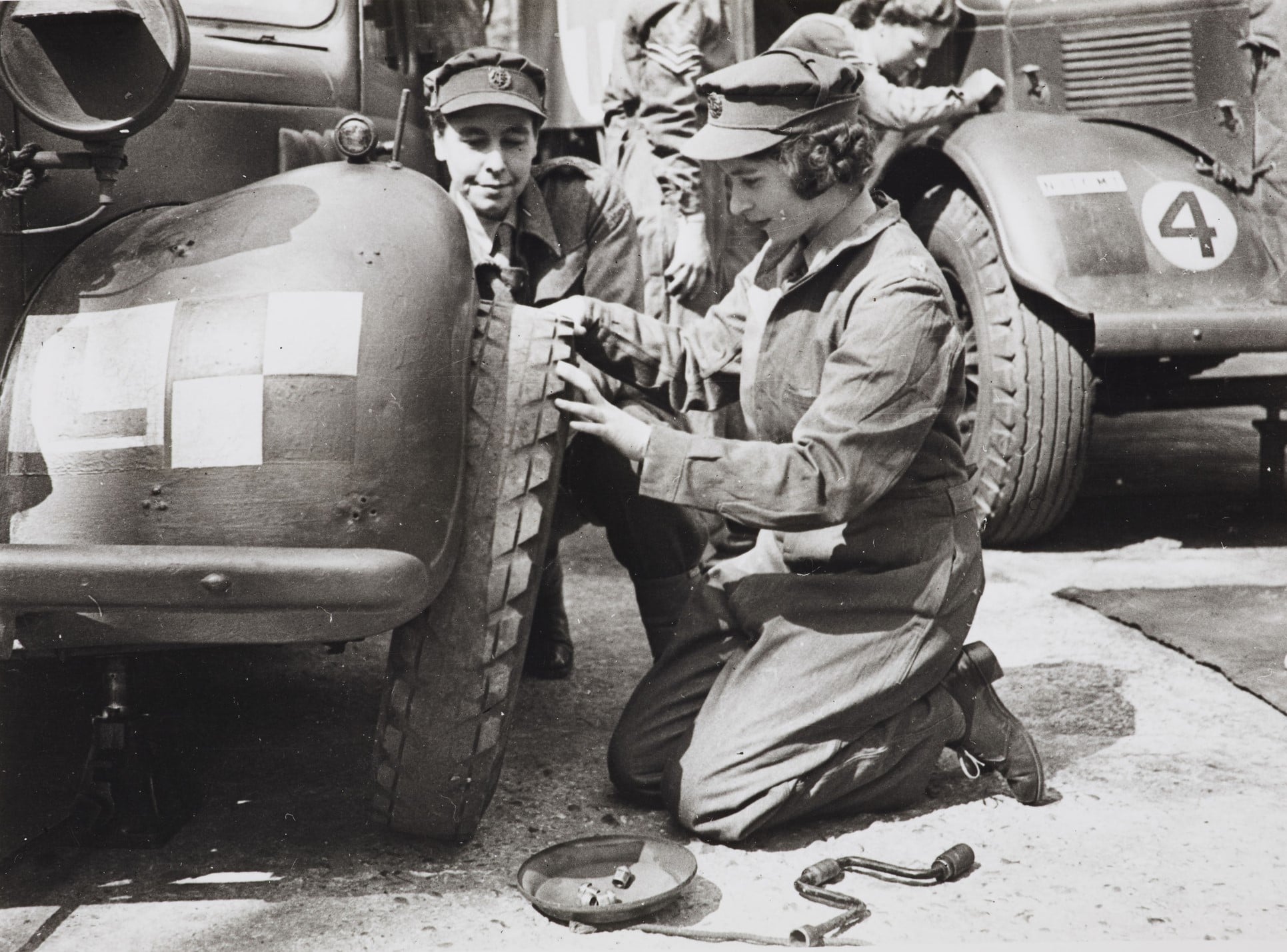 Princess Elizabeth trains as an A.T.S Officer at a Training Centre in Camberley, Surrey April 1945 (RCIN 2002237) Photograph of Queen Elizabeth II when Princess Elizabeth, wearing overalls, kneeling down facing left, as she takes the right front wheel off a car during a Motor Vehicle Maintenance training.