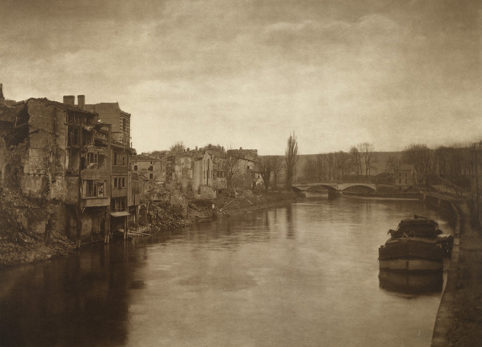 Olive Edis, Verdun, 1919 (RCIN 2943161) Photograph of a view of the River Meuse, Verdun, by Olive Edis. On the left bank are a series of ruined buildings, surrounded by debris. On right bank is a towpath and a moored small barge. A bridge surrounded by trees is visible in the background.