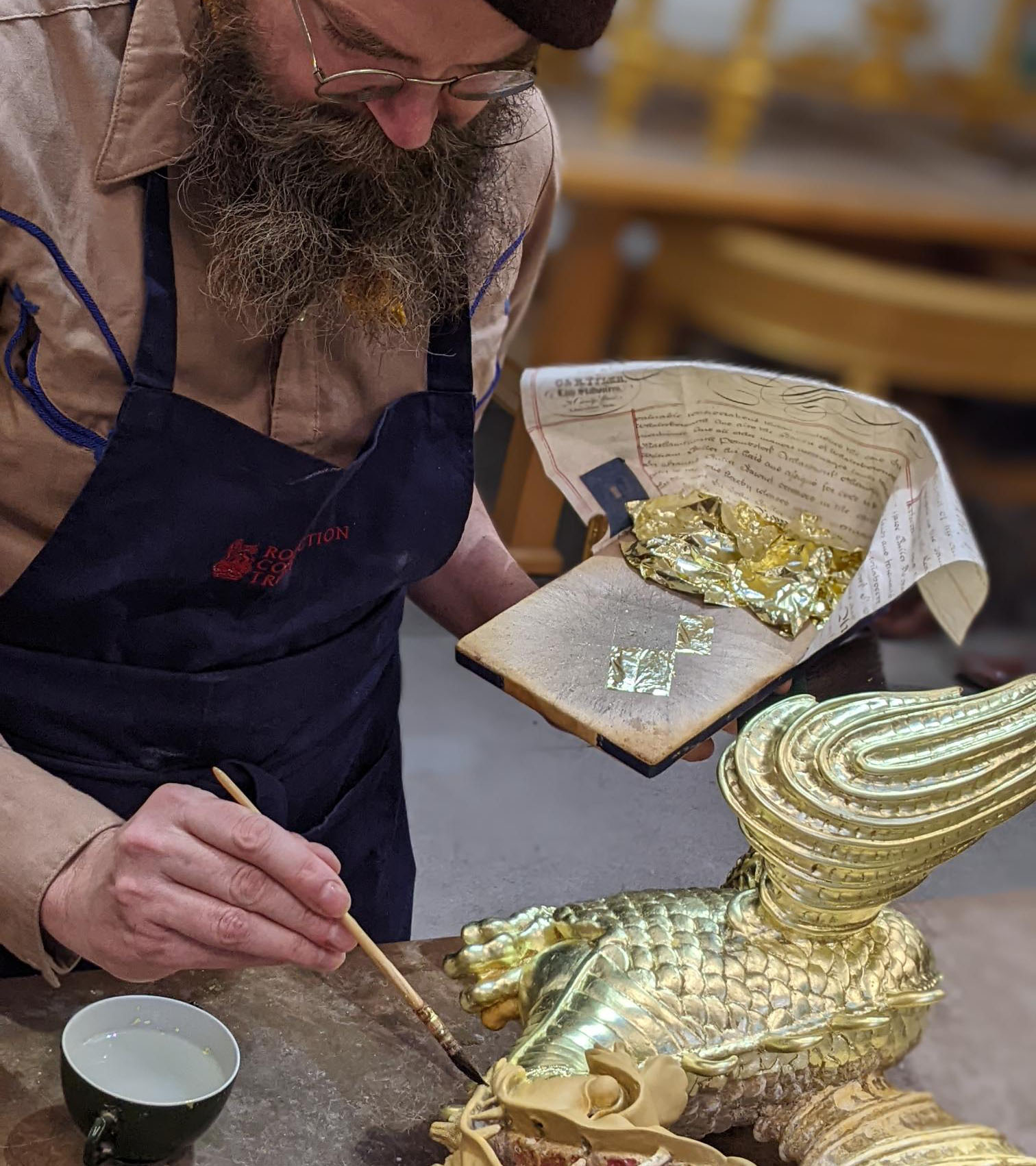 A conservator adding gold to one of the dragons A standing man holds a paintbrush with gold leaf onto a dragon object.