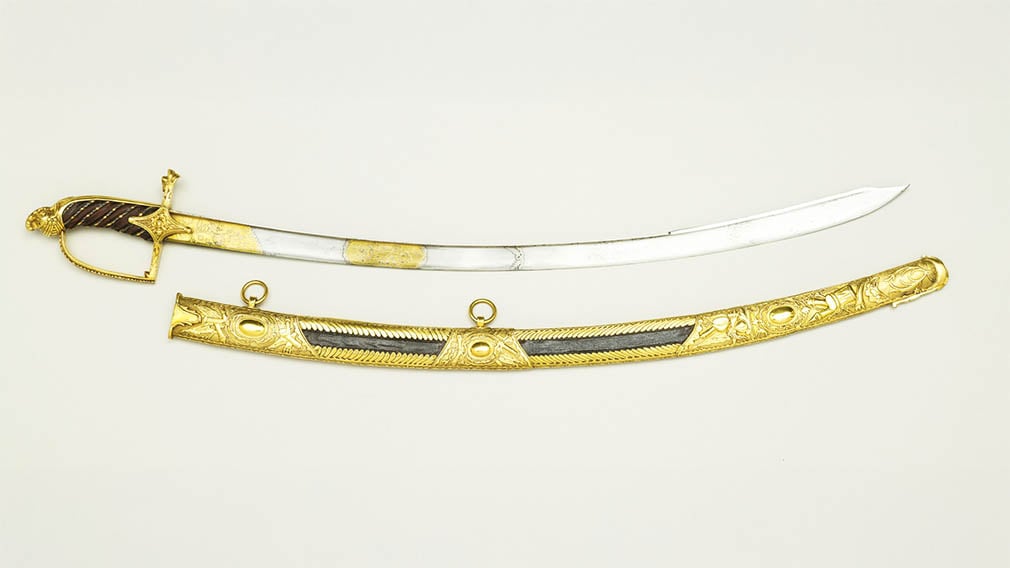 Prussia, Sabre and scabbard 1805-10 (RCIN 61140) Ornate stirrup-hilted sabre with a gilt metal hilt, featuring a knuckle-guard adorned with foliage and acorns, a quillon block chased with trophy designs, and a leather-wrapped grip shaped like a helmet.