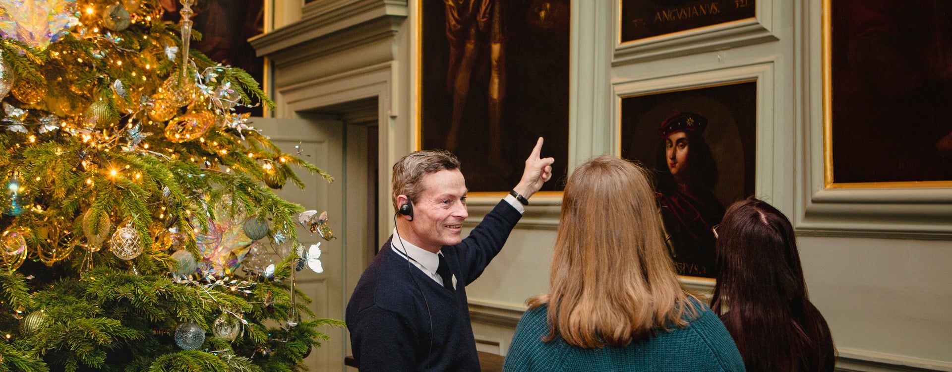 Christmas Descriptive Tour A man pointing at a painting. Two other people look at the painting on the wall. A Christmas tree is next to them.