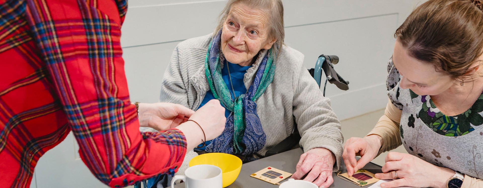 Two people including an older woman in a wheelchair are crafting at a table.