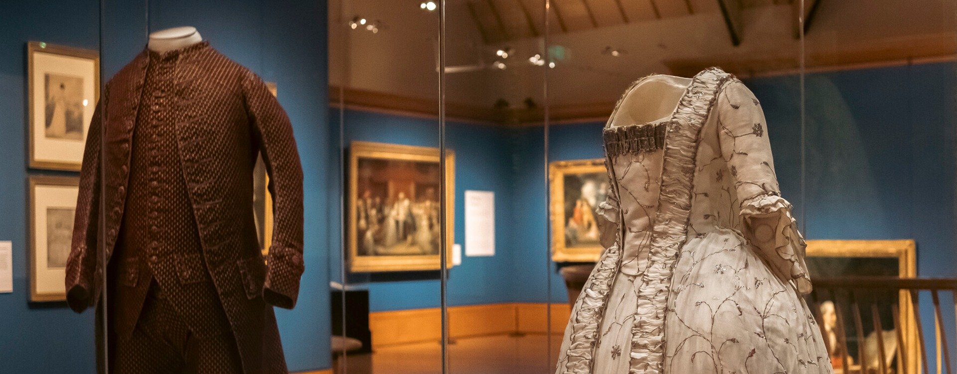 A historic dress and suit in a glass cabinet with paintings visible behind