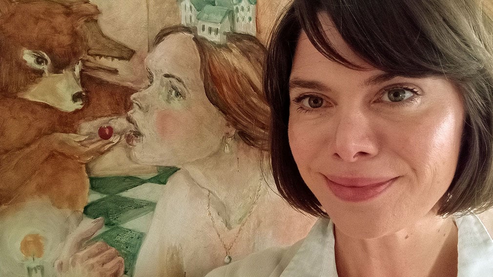 A woman with short brown hair and white top stands in front of a painting